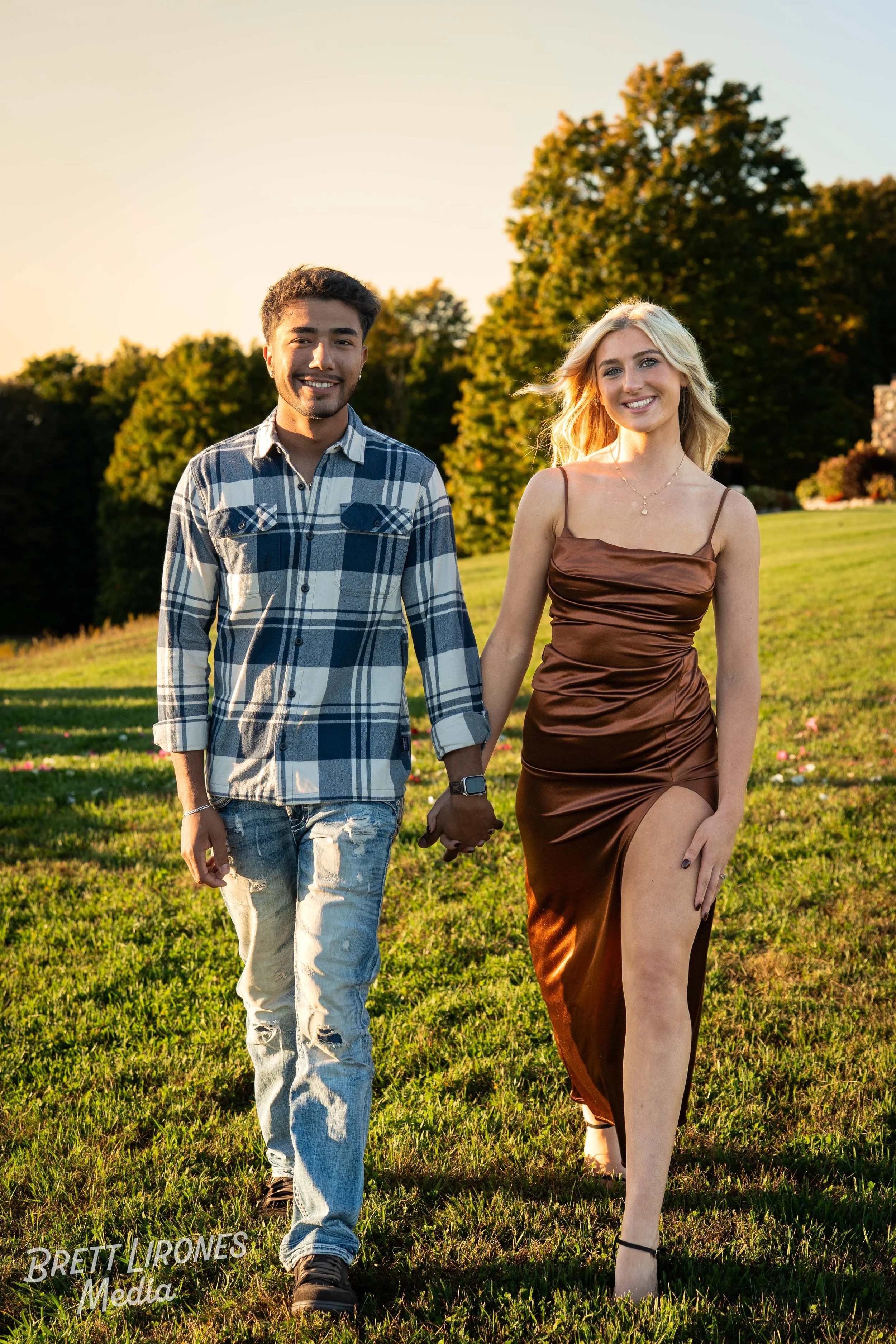 A young couple holding hands and walking in a grassy field during sunset, with trees in the background.