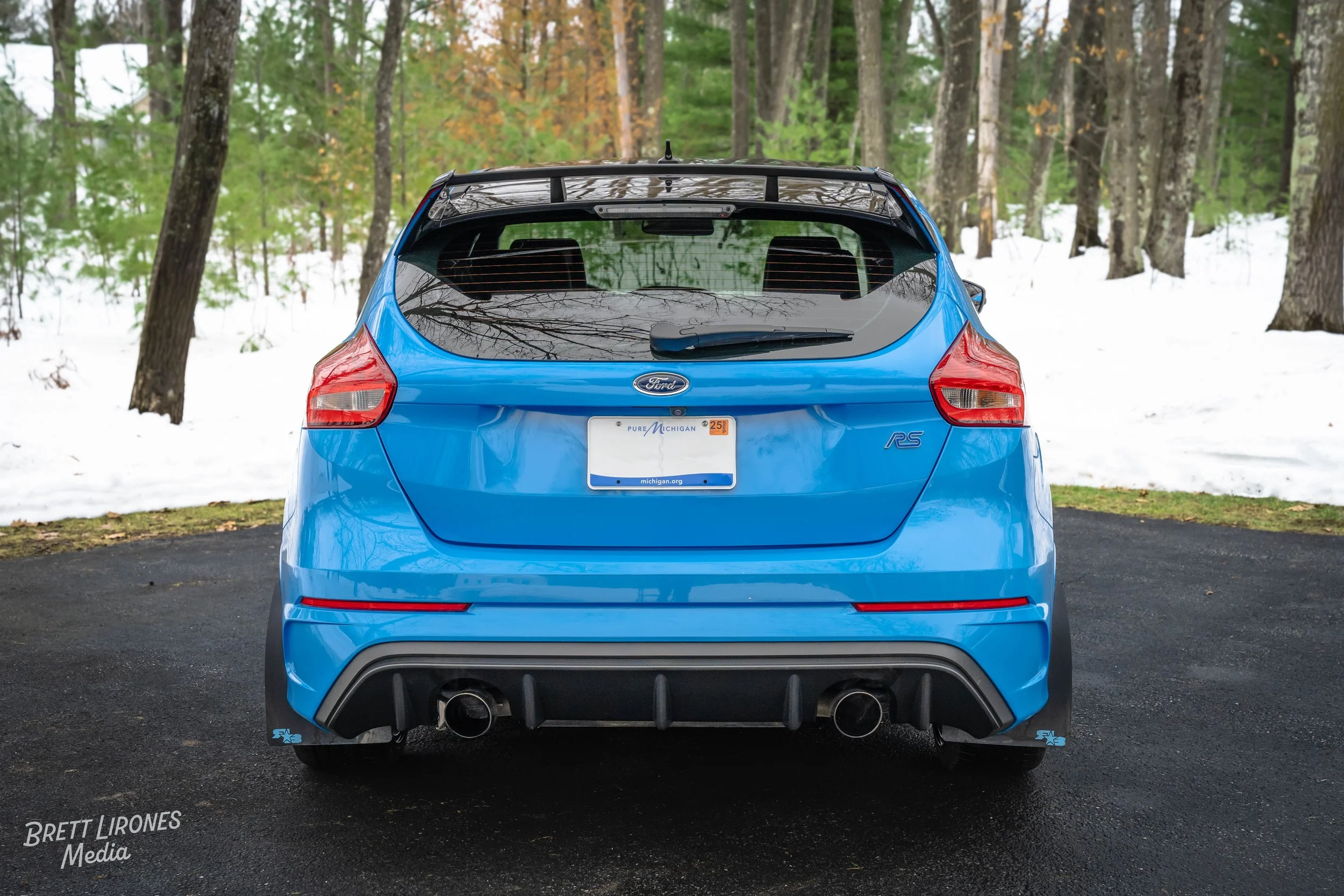Rear view of a blue Ford Focus RS car parked on a black road with snow and trees in the background.