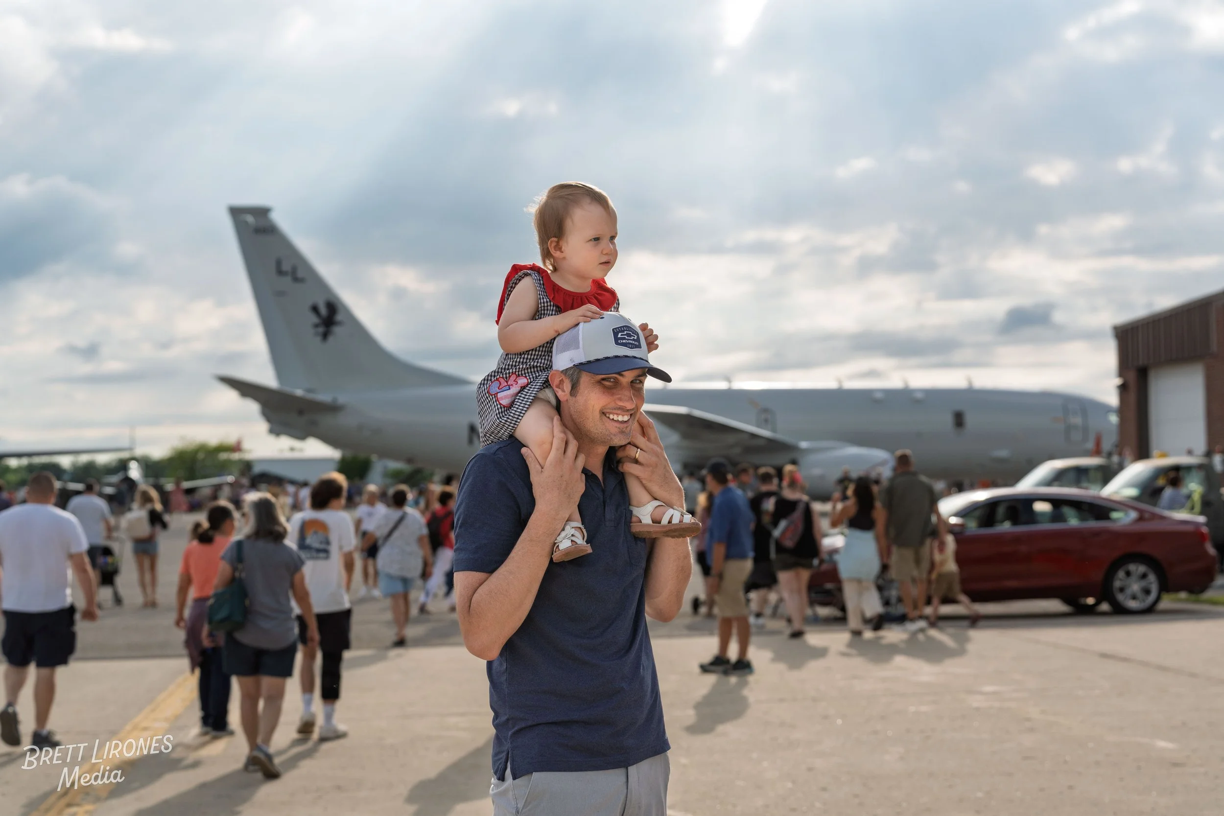 A man carrying a young girl on his shoulders at an airshow, with a large military aircraft and a crowd in the background.