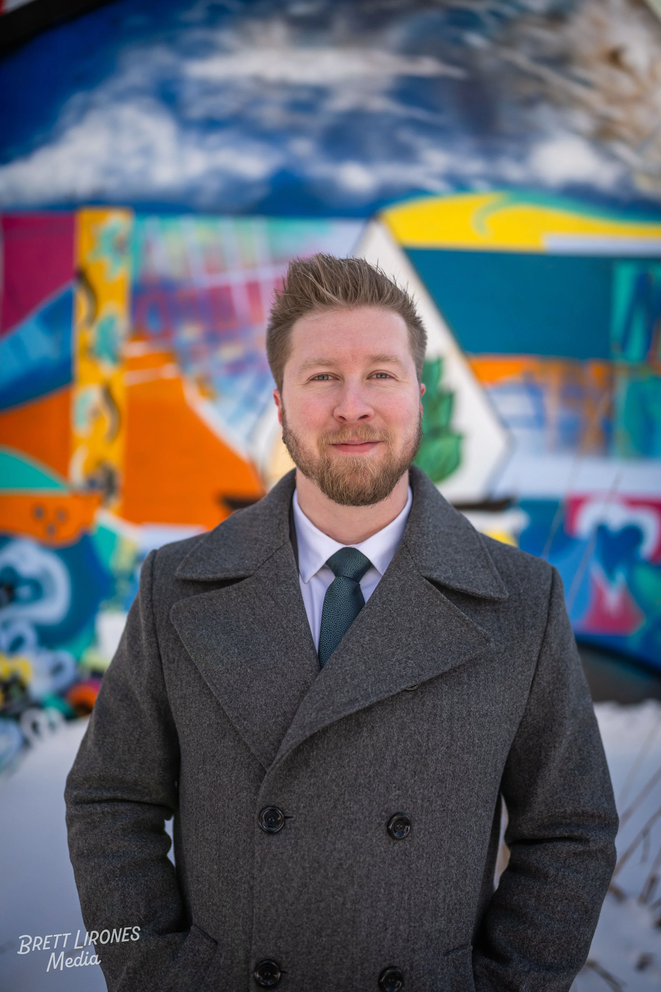 A man with a beard wearing a gray coat and tie standing outdoors in front of a colorful graffiti wall
