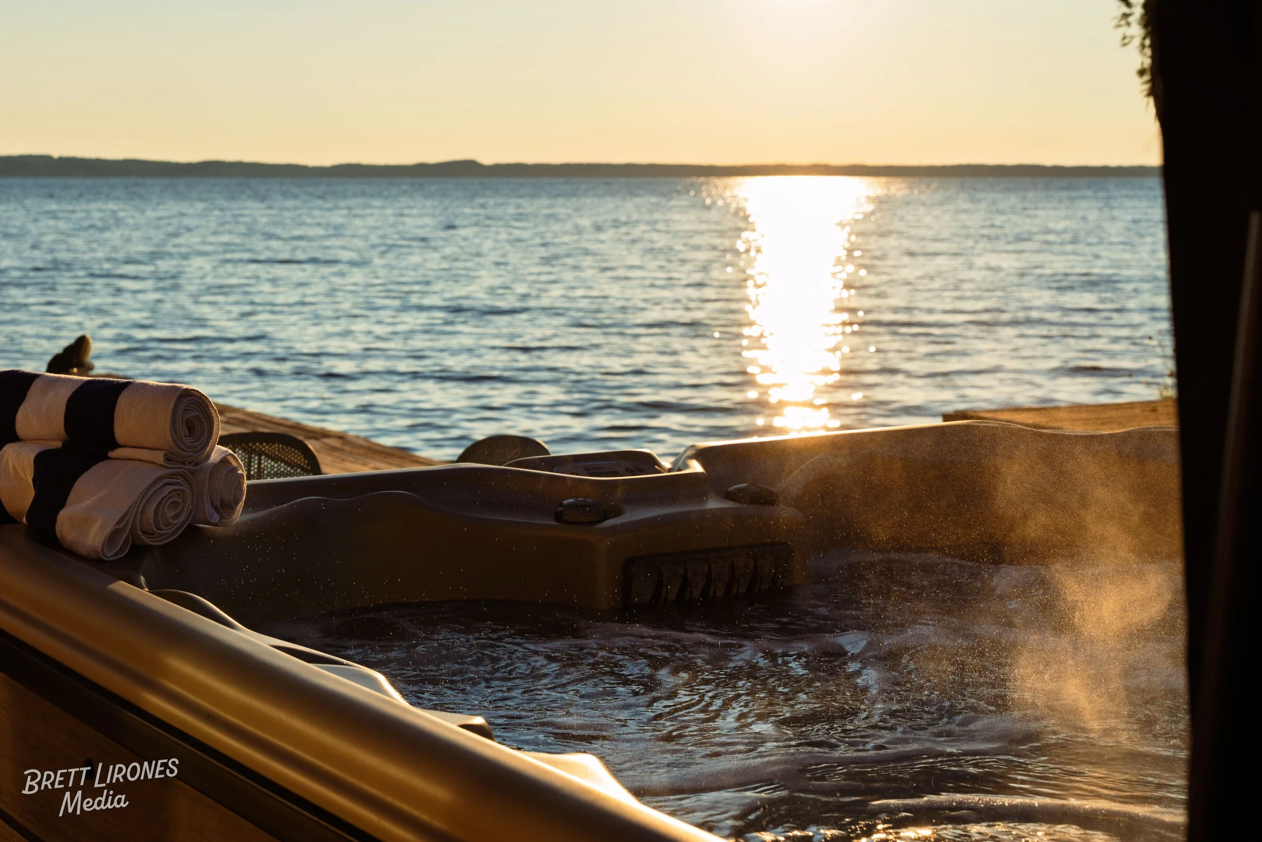 A hot tub on a dock by the water at sunset with rolled towels on the edge.