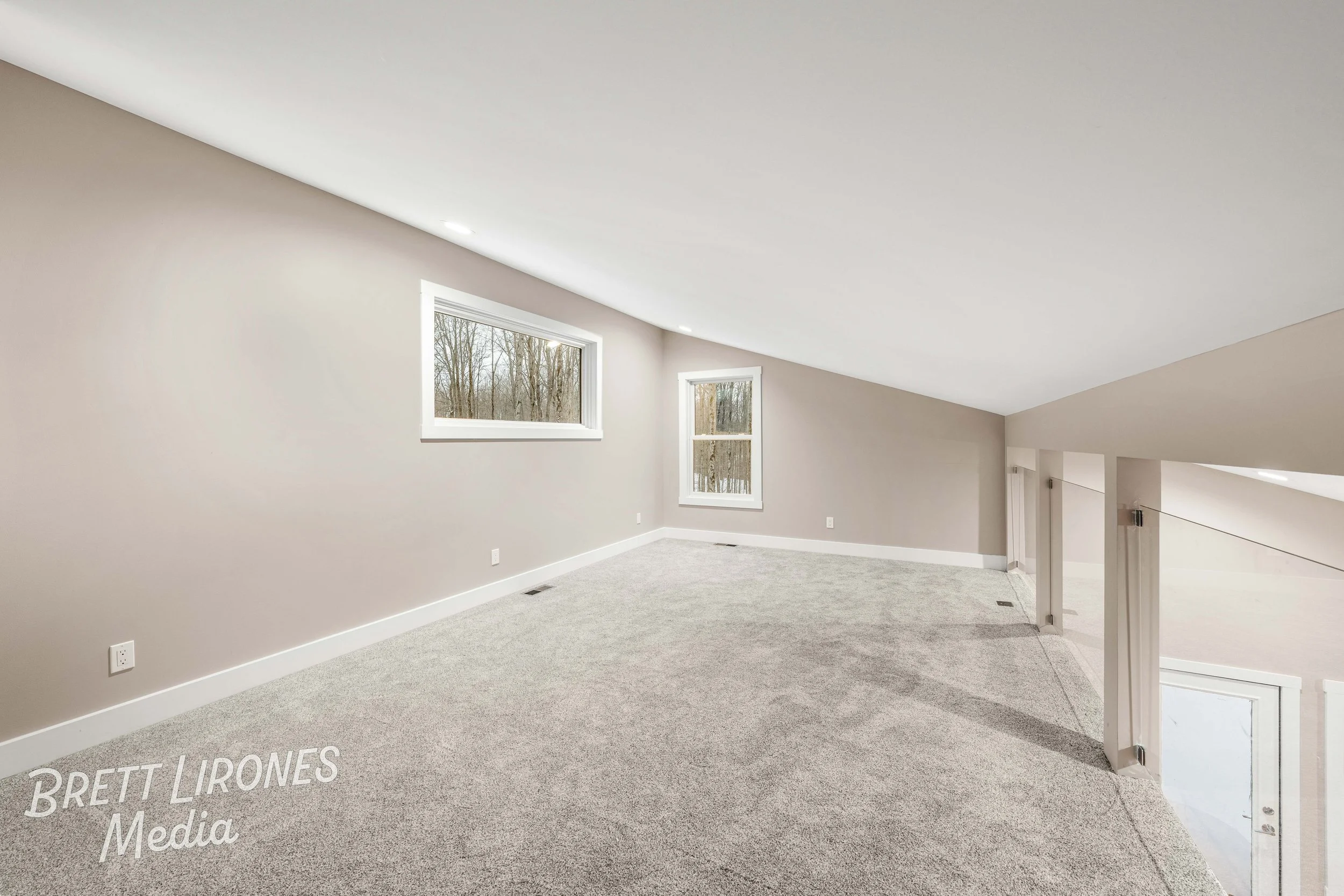 Empty finished attic space with two windows, beige walls, light gray carpet, and white trim, overlooking a tree-lined area outside.