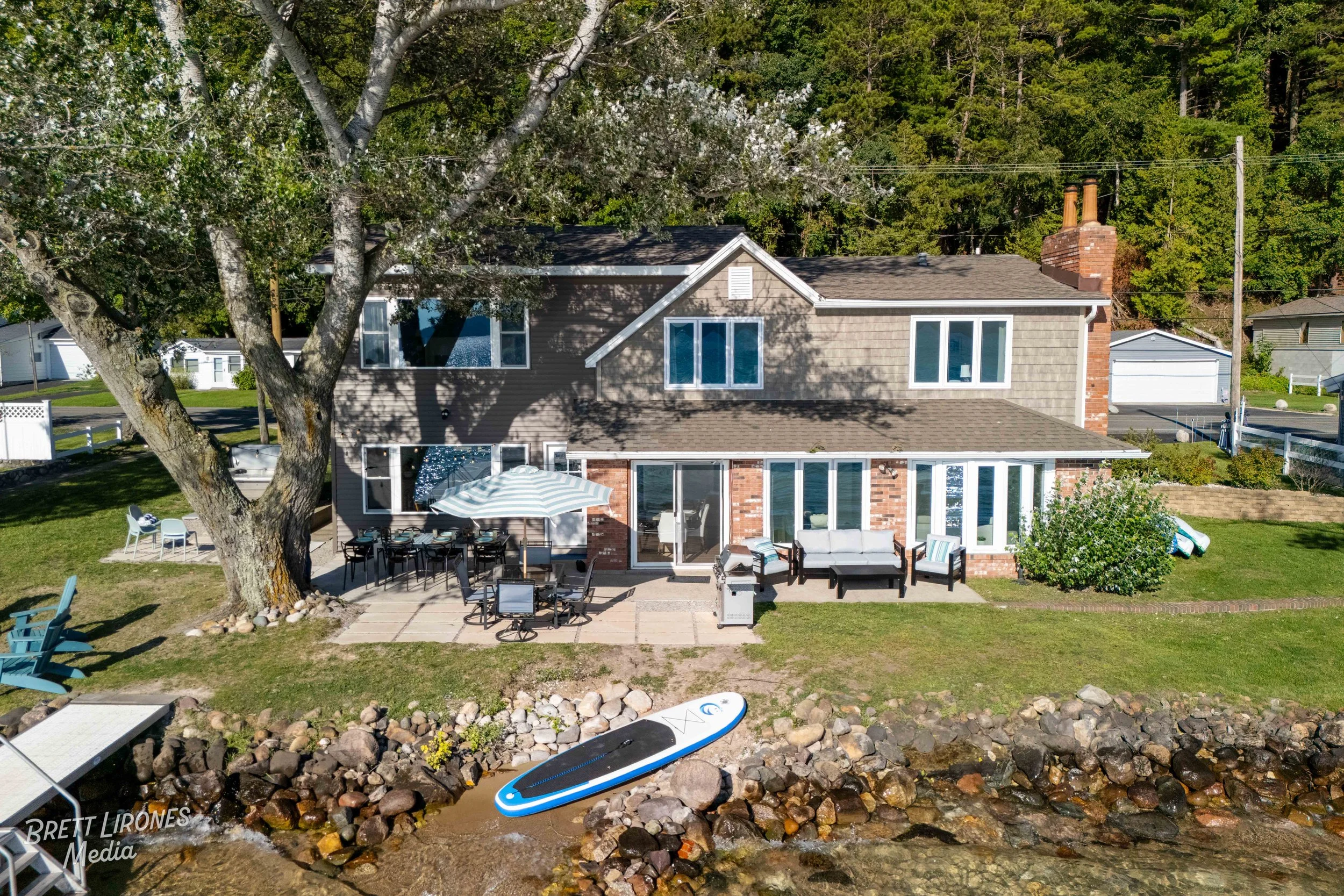 View of a two-story house with a backyard patio, large tree, outdoor furniture, a paddleboard on the rocks near the water, and a dock, with wooded area in the background.