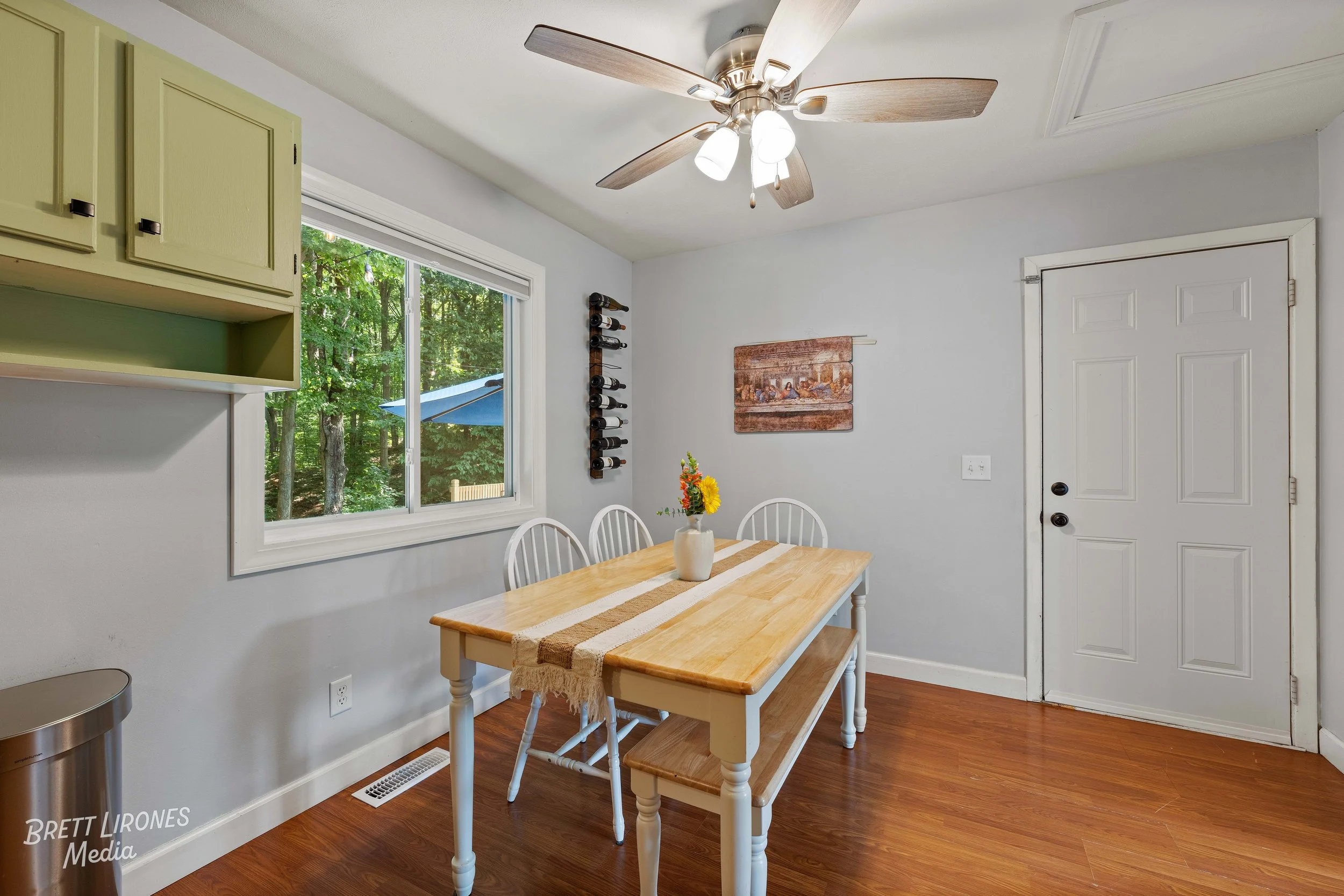 Dining room with a wooden table, white chairs, a flower vase, and a window showing green trees outside. There is a ceiling fan with lights and a wall-mounted wine rack.