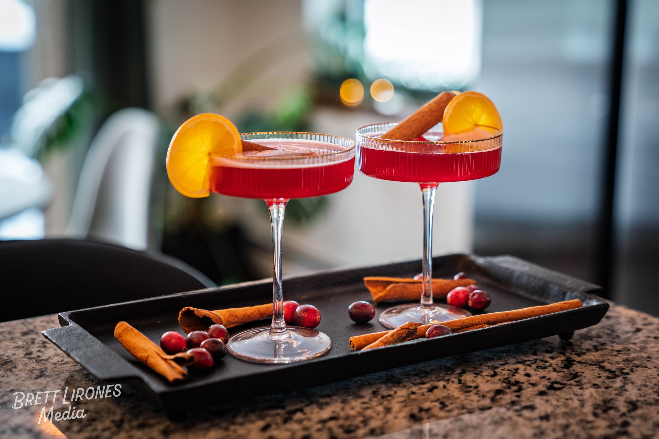 Two glasses of red cocktail garnished with orange slices and cinnamon sticks, placed on a black tray with cranberries and cinnamon sticks on a granite countertop.