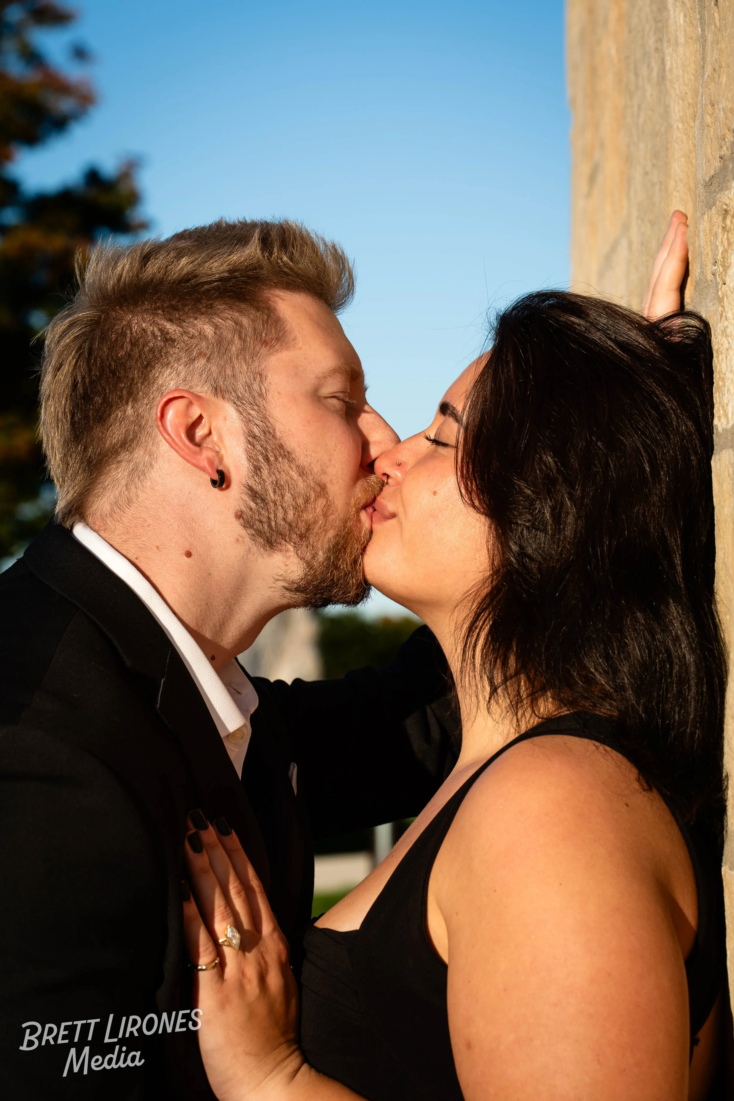 A couple is kissing outdoors, with the woman holding the man's chest and her back against a wall, during daytime with a clear blue sky.