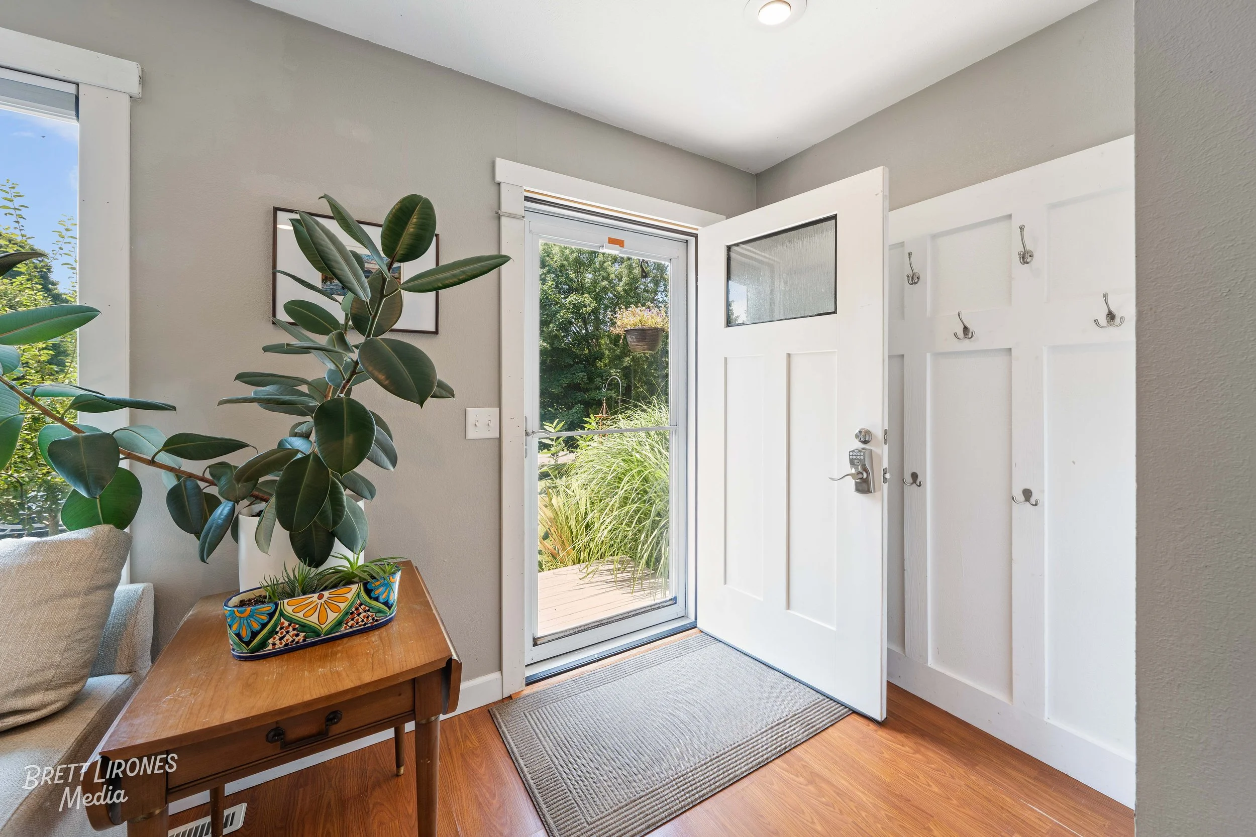 Interior view of a front door area with a white door and a window, a wooden side table with a potted plant, a gray rug, and a wall-mounted coat rack with hooks. Outdoors, greenery is visible through the door and window.