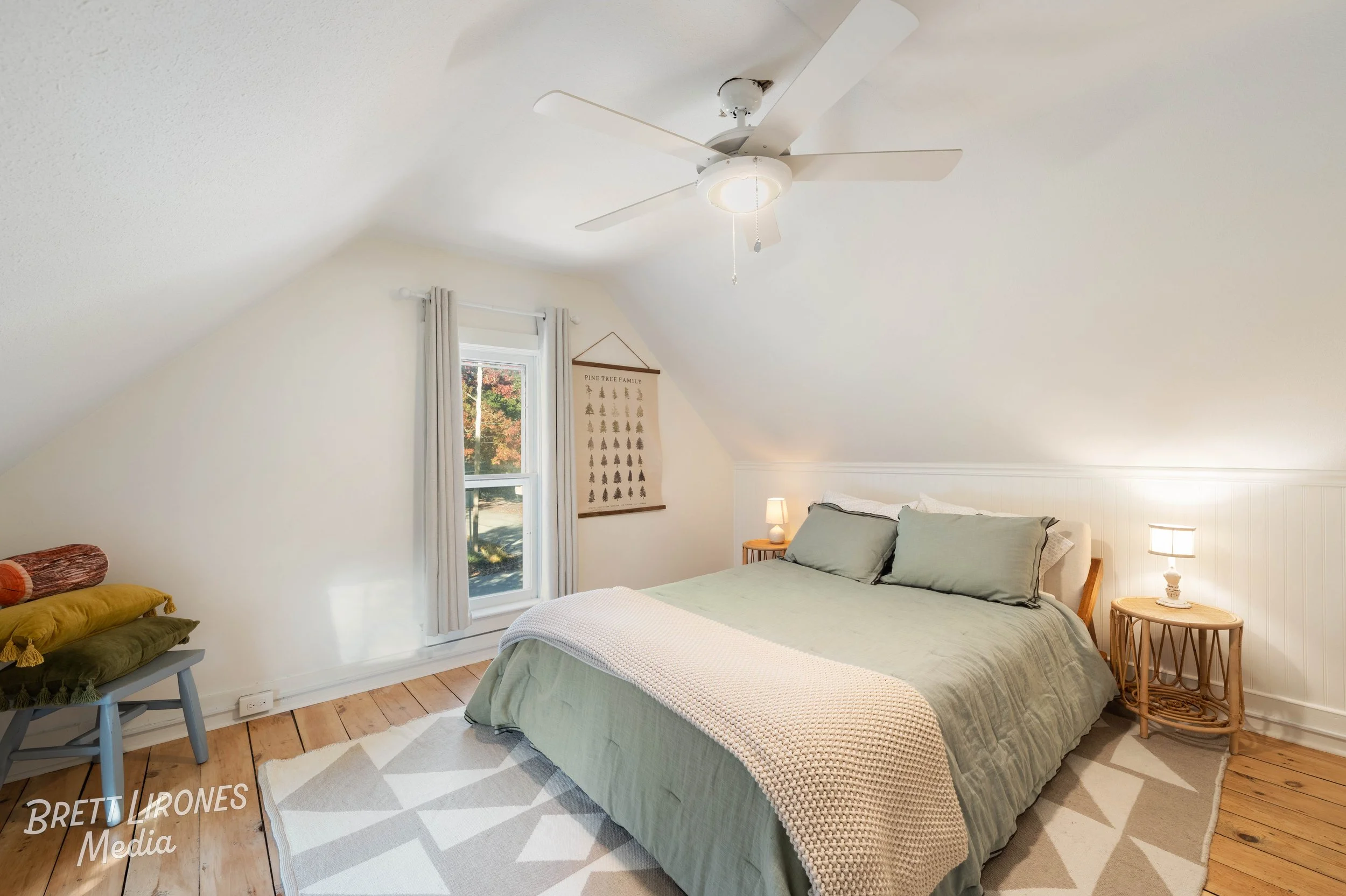 A bedroom with a sloped ceiling, a bed with light green bedding, two wooden nightstands with lamps, a window with white curtains, a ceiling fan, and a wall hanging with tree illustrations.