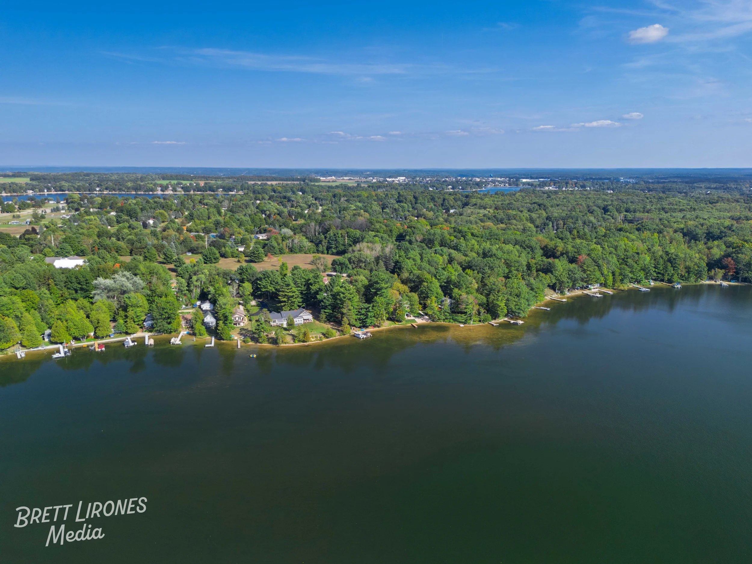 Aerial view of a lakeside neighborhood with trees, houses, and docks along the shoreline, under a partly cloudy blue sky.