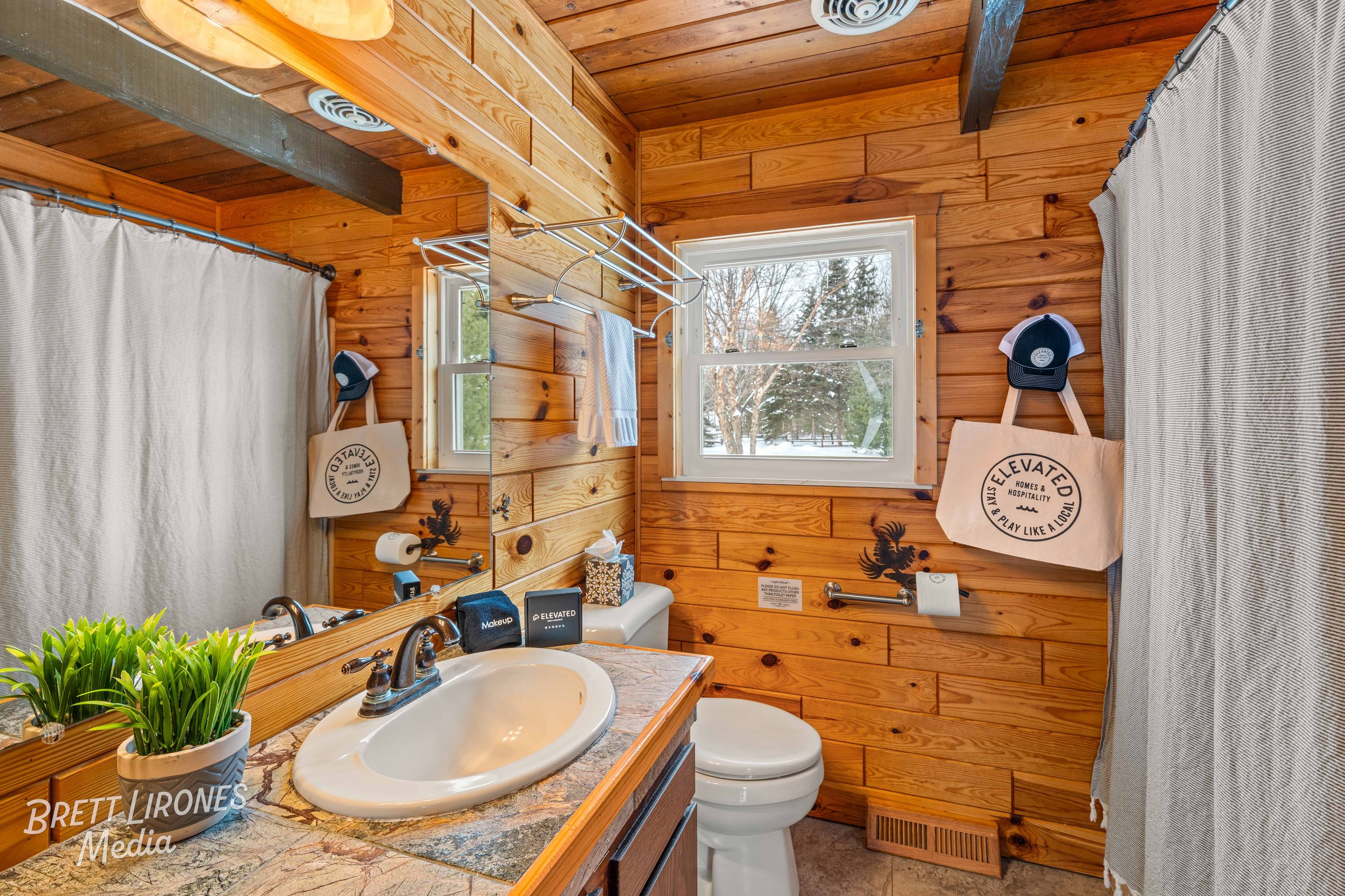 Wood-paneled bathroom with a white sink, a small mirror, a window showing outdoor trees with snow, and various personal items and decor.