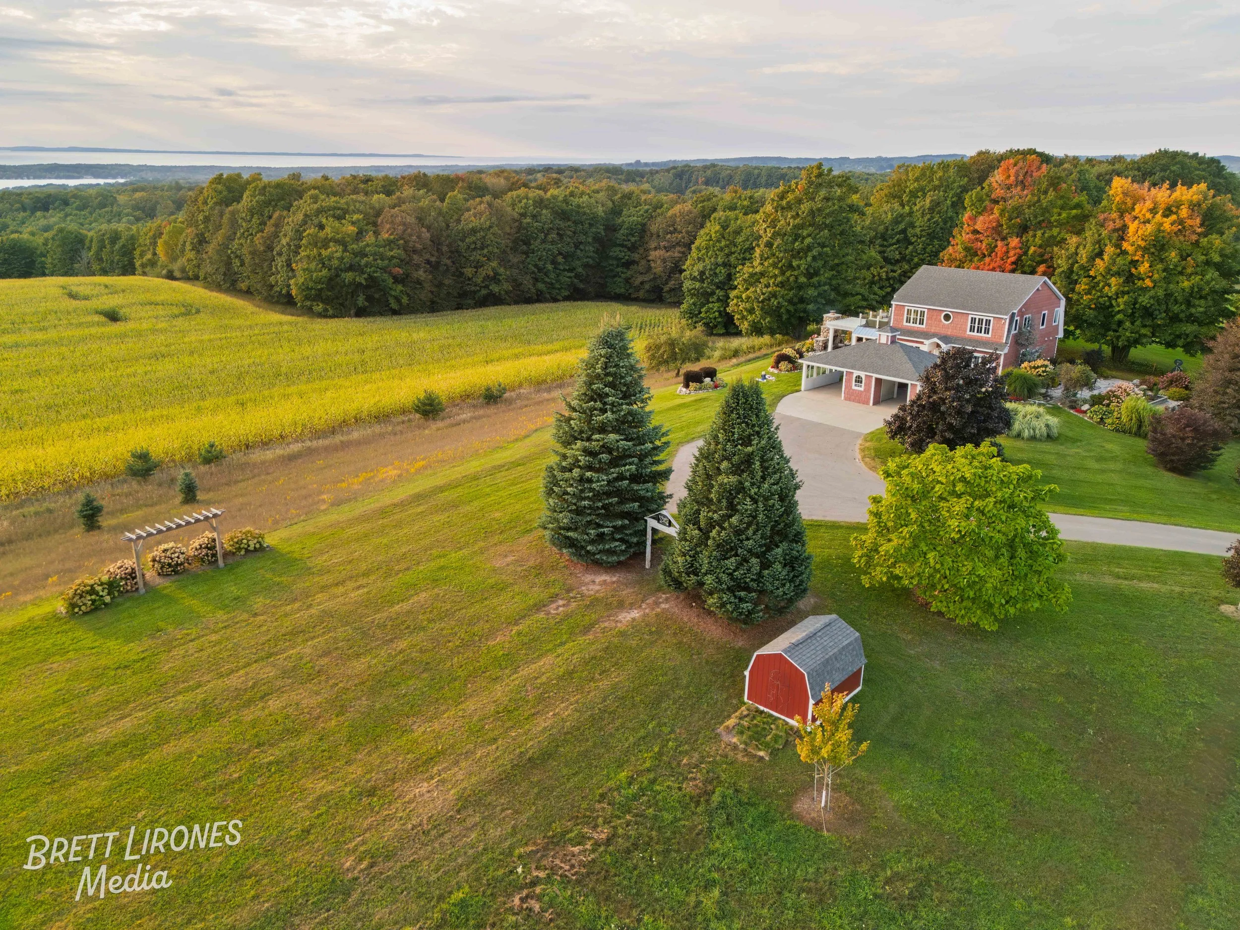 Aerial view of a large house with a driveway and a red barn on a lush green property surrounded by trees, fields, and a distant body of water under a cloudy sky.