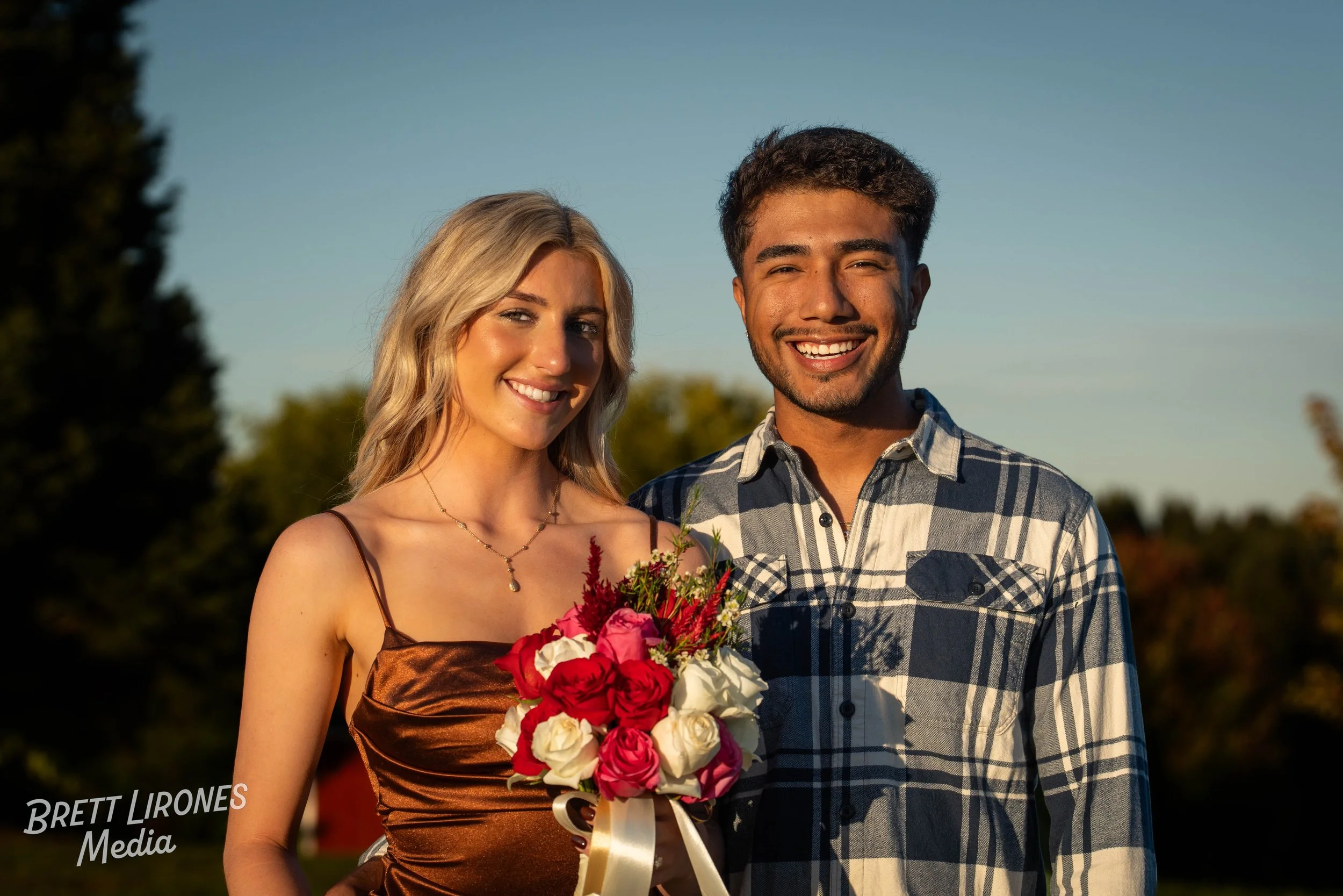 A young woman and man stand outdoors smiling, with the woman holding a bouquet of roses, during sunset.