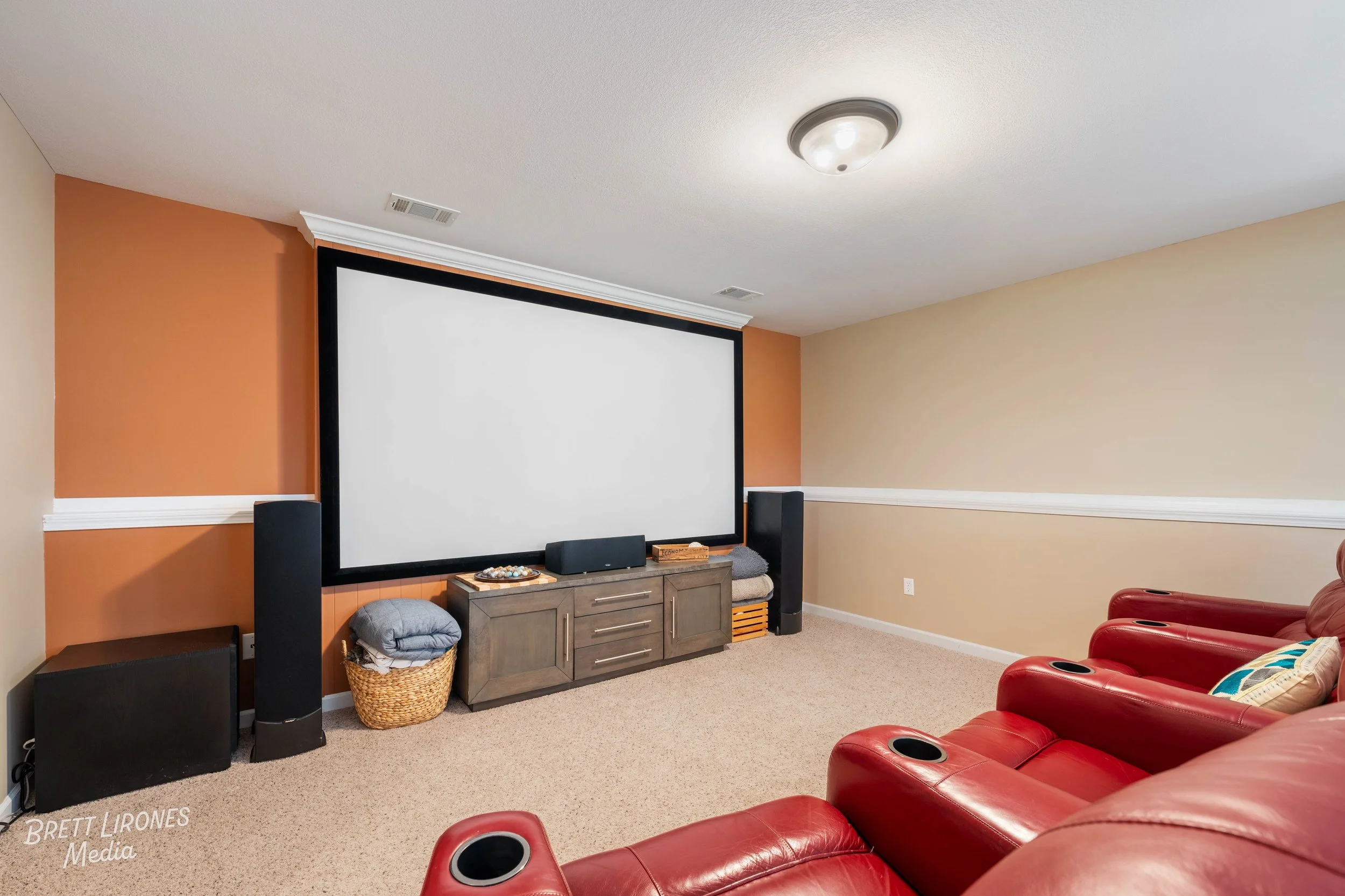 A home theater room with a large projector screen on an orange wall, red leather recliners, a wooden cabinet with electronic equipment, and carpeted floor.