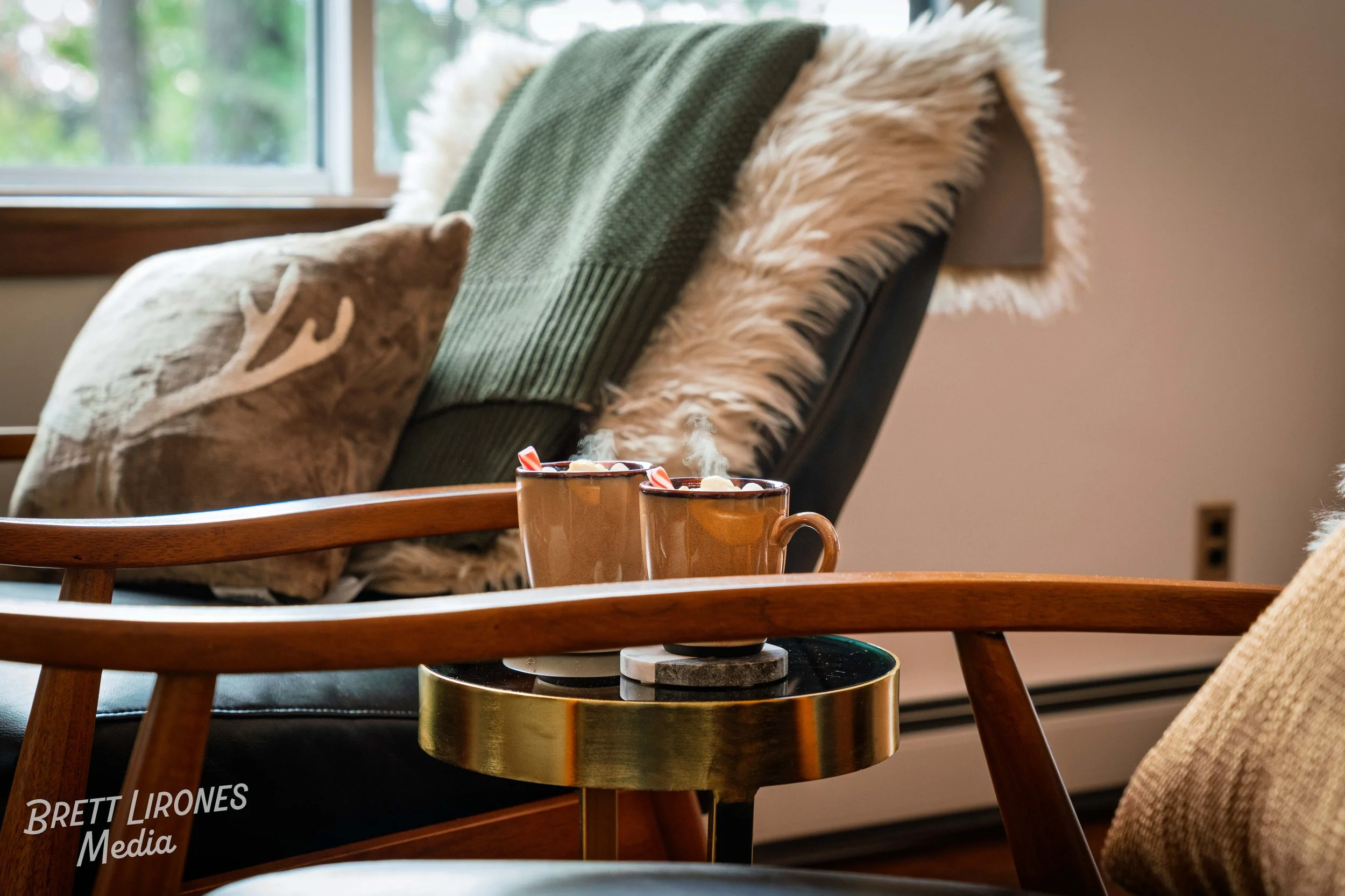 Two steaming mugs on a round side table in a cozy living room, with armchair and cushions nearby.