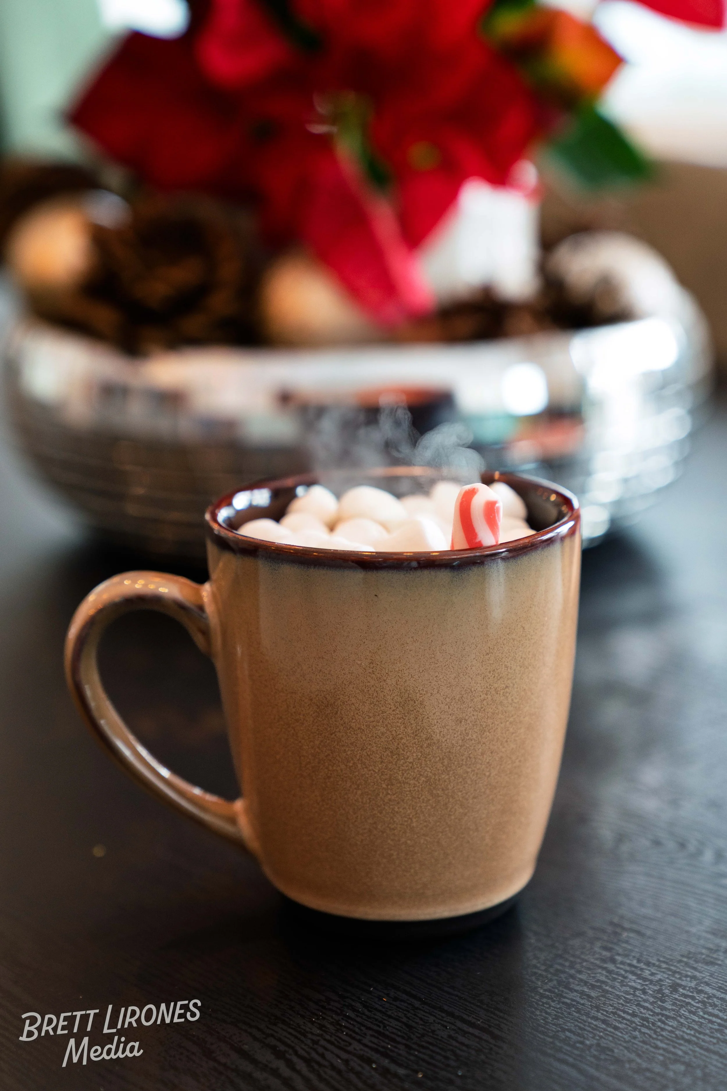 A beige mug filled with hot chocolate topped with mini marshmallows and a small candy cane. In the background, a silver tray holds Christmas decorations, including a poinsettia and pinecones.