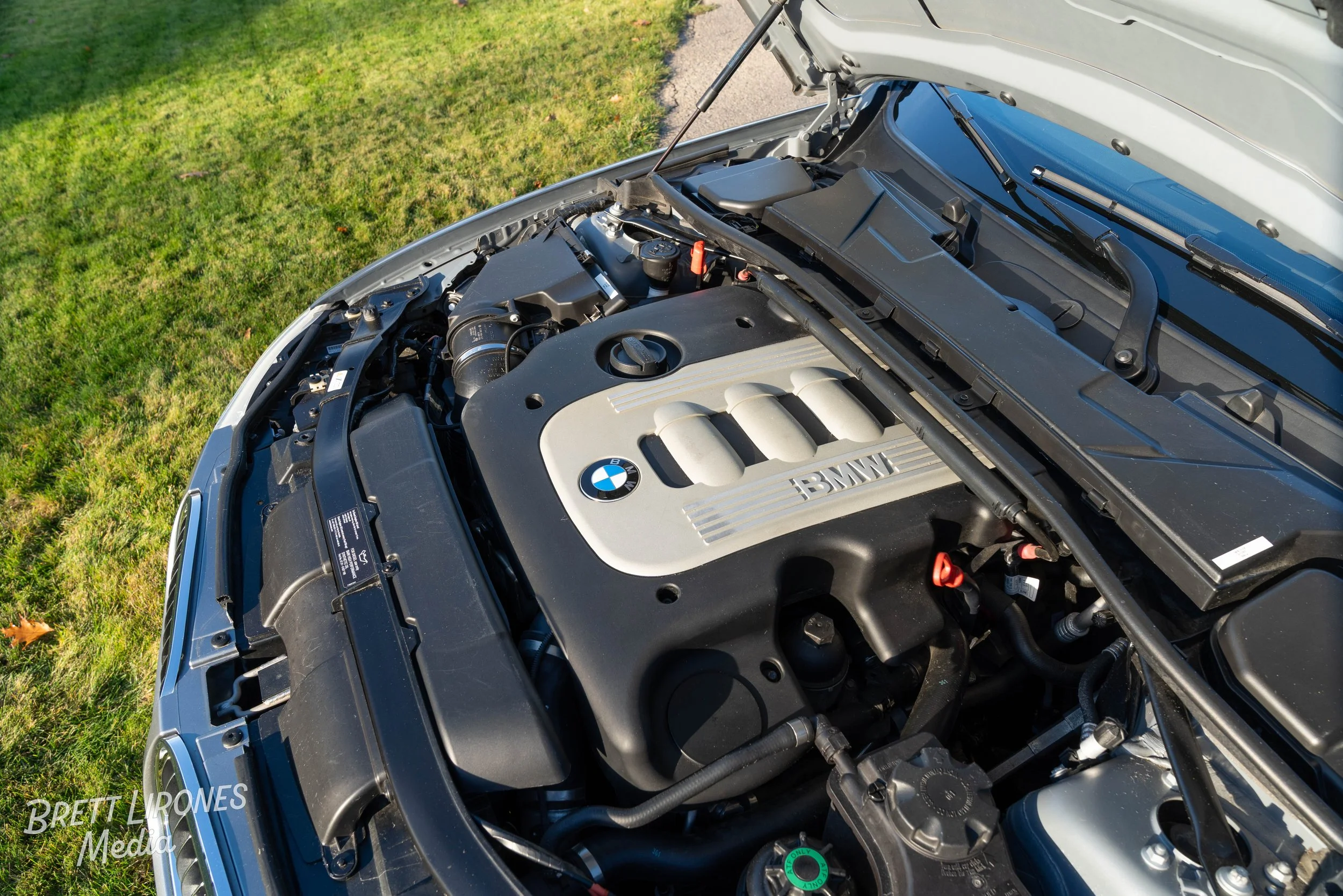 Open hood of a BMW car showing the engine compartment with a silver and black engine cover bearing the BMW logo. The car is parked on grass.