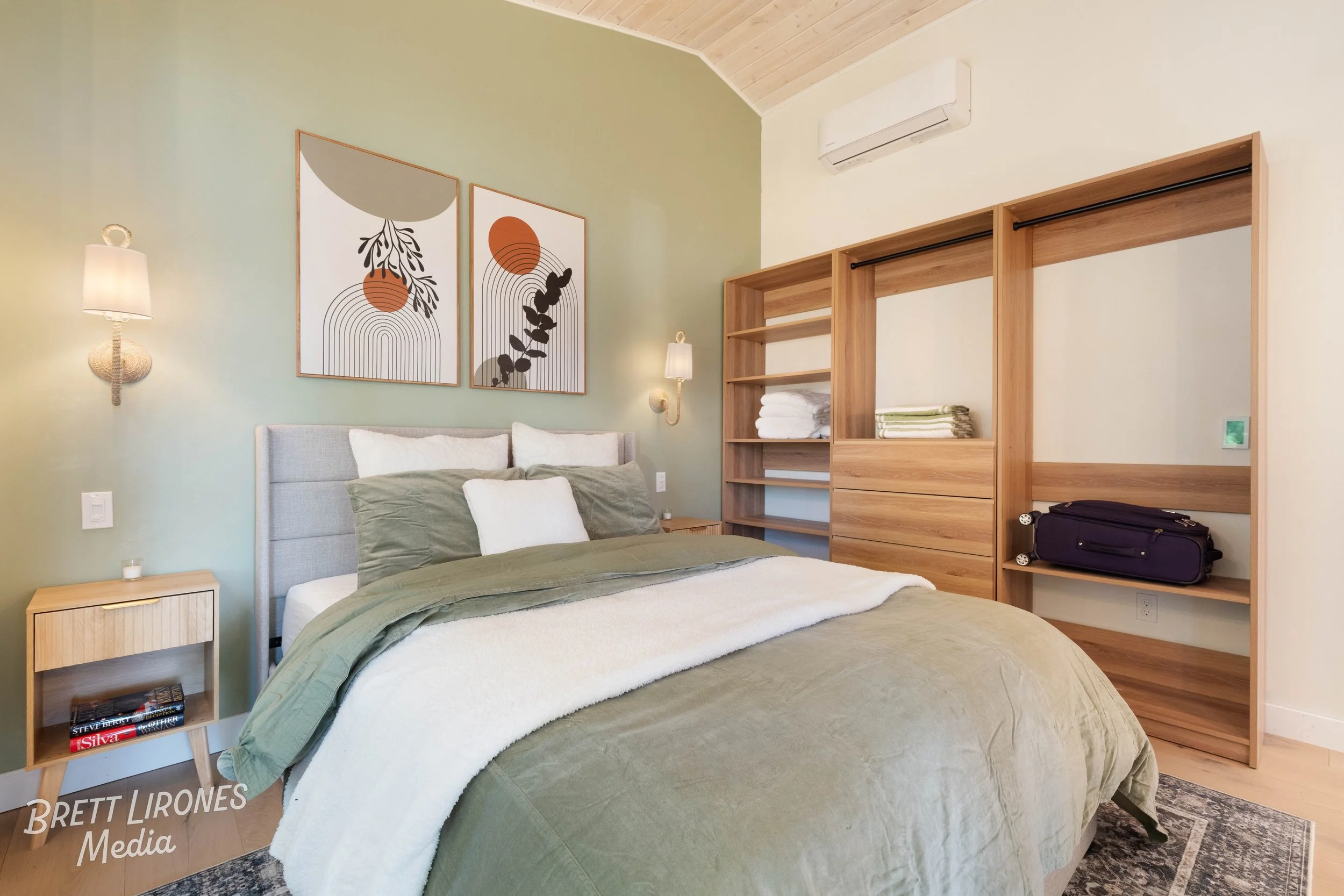 A bedroom with a bed featuring green, white, and beige bedding, a wooden nightstand with books, and artwork above the bed. There are built-in wooden shelves with folded towels and a purple bag, wall-mounted lamps, and a wall-mounted air conditioner.