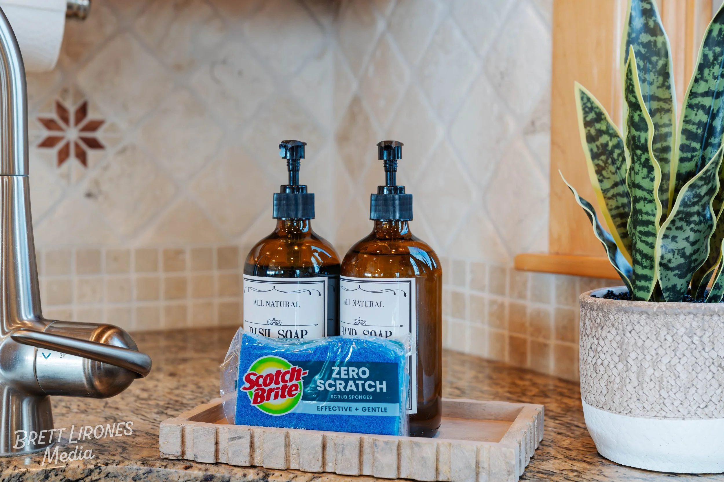 A kitchen counter with two amber soap dispenser bottles labeled 'All Natural Dish Soap' and 'Hand Soap', a blue Scotch-Brite scrub sponge labeled 'Zero Scratch', a potted snake plant in a textured white pot, and a chrome kitchen faucet in the backgro