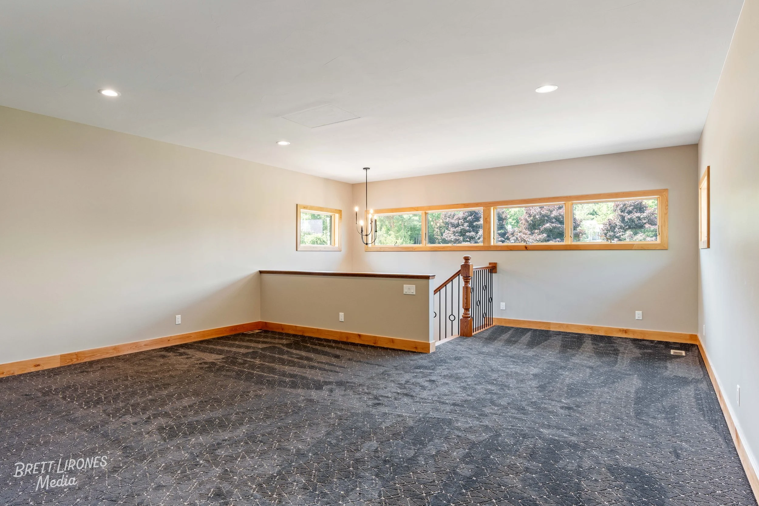 Empty living room with beige walls, multiple small windows, dark patterned carpet, wooden trim, chandelier, and staircase with wooden banister and black iron balusters.