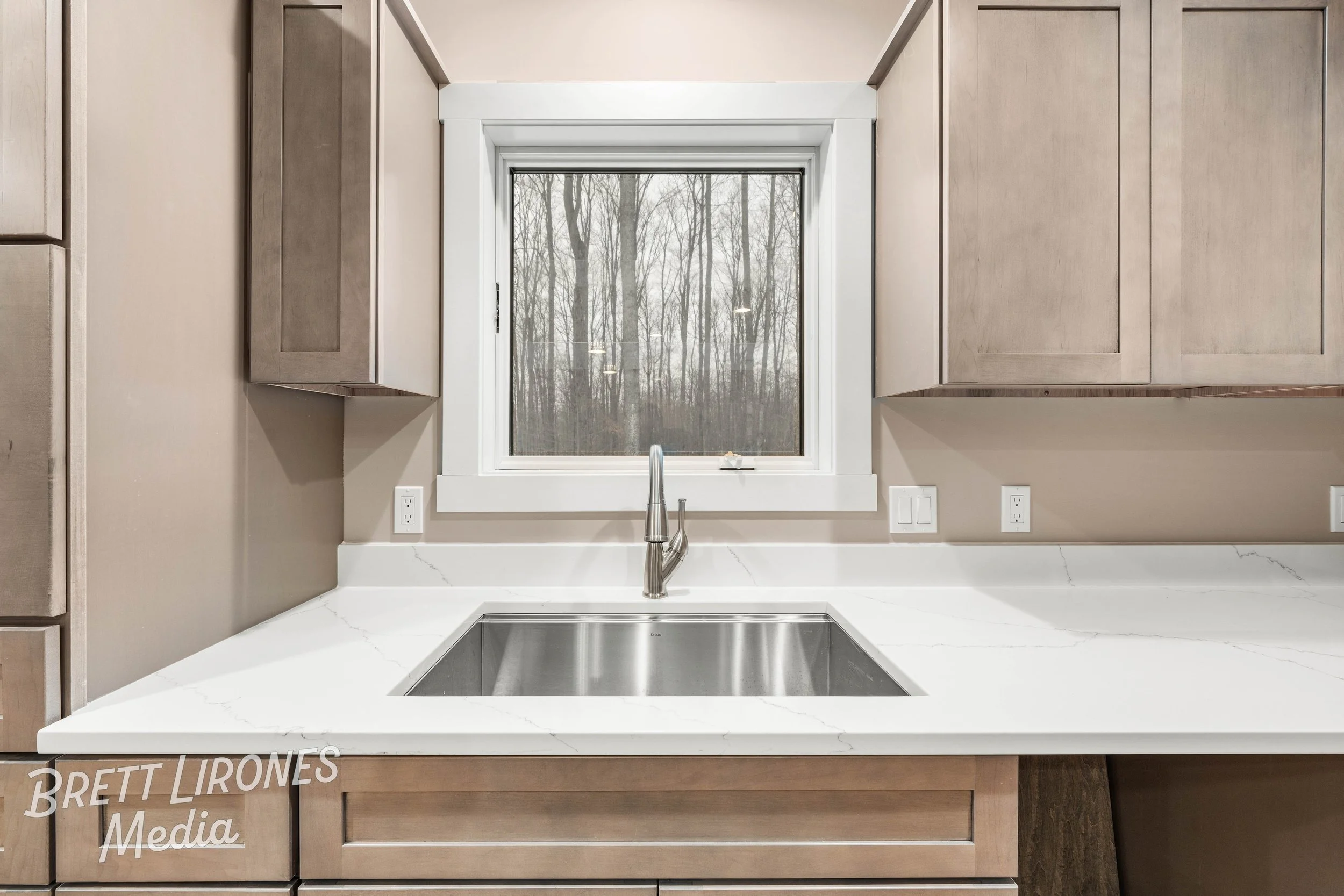 Kitchen counter with a stainless steel sink, a window above, beige cabinets, and a view of leafless trees outside.
