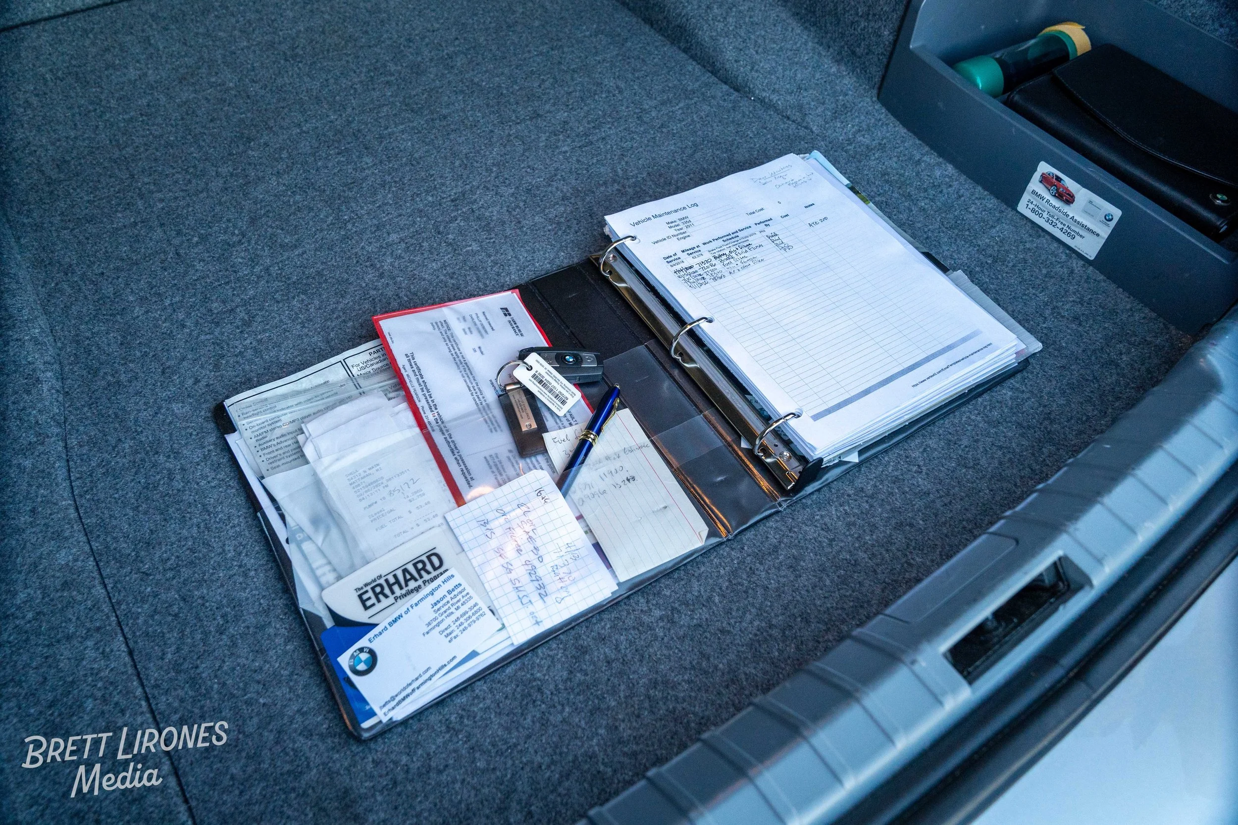 Car trunk with a folder containing vehicle maintenance records, business cards, a pen, and receipts on a carpeted surface.