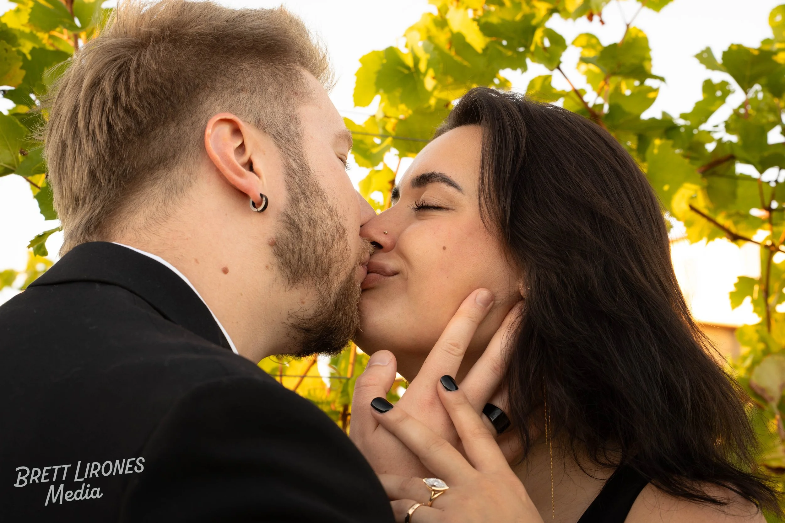 A couple is kissing outdoors with green and yellow leaves in the background.