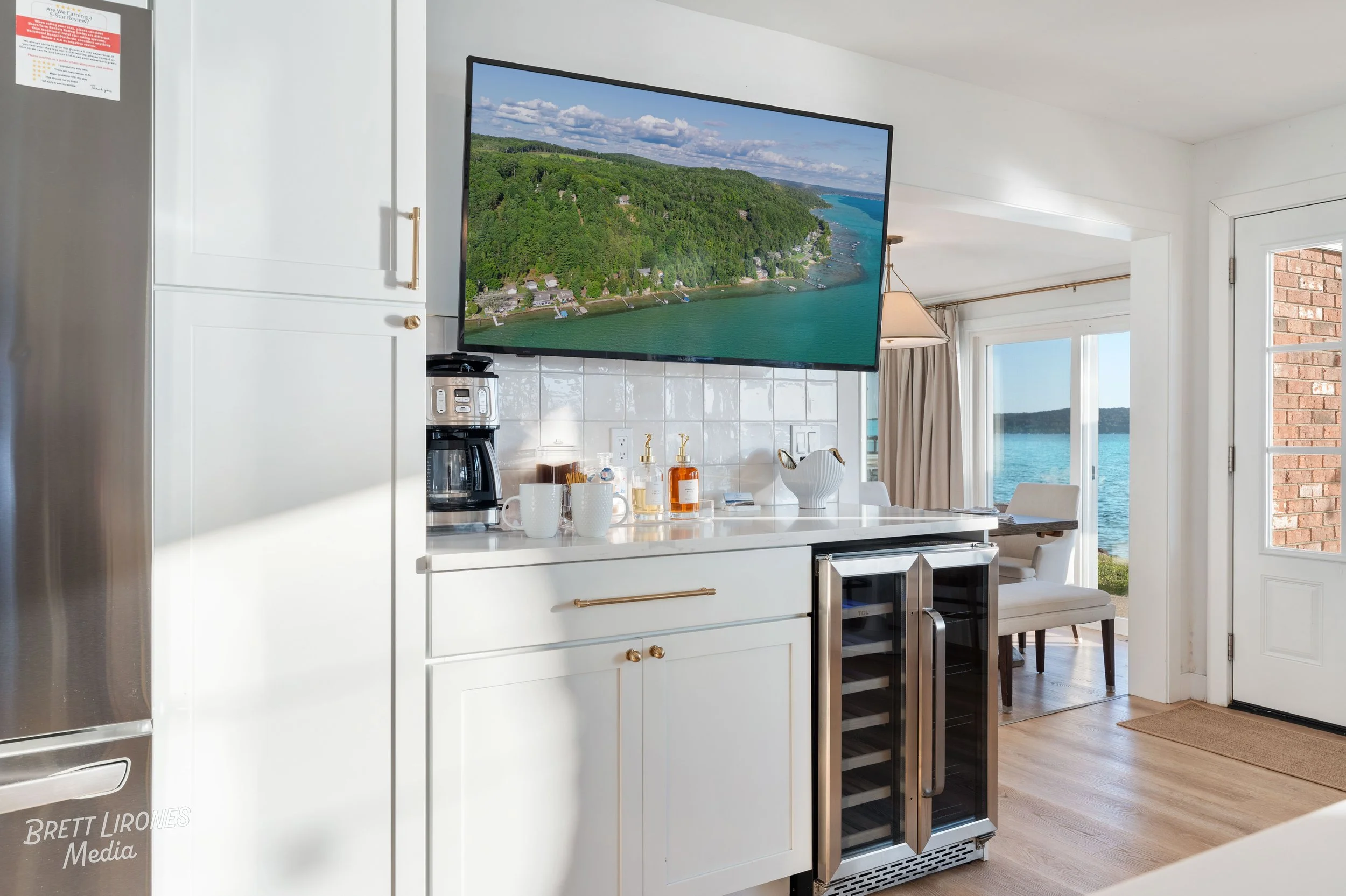Kitchen with white cabinets, a coffee maker, mugs, soap dispensers, a small wine fridge, and a wall-mounted TV displaying an aerial view of a lake and green hills, with a dining area and waterfront view visible through glass doors.