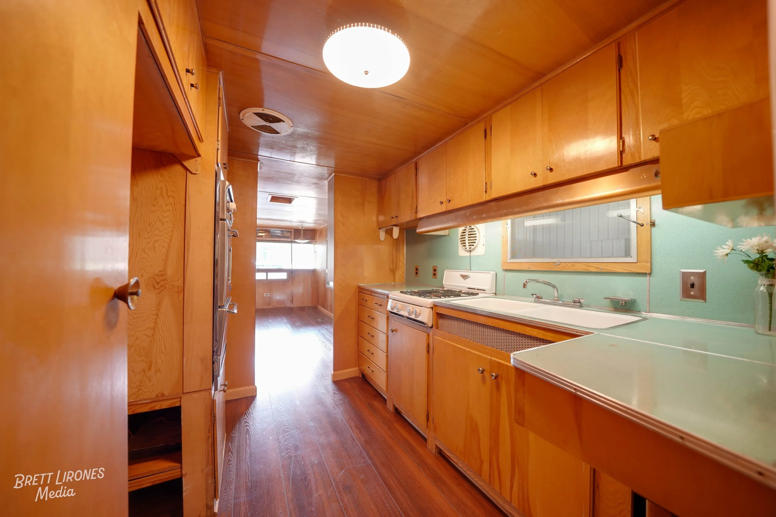 Kitchen with wooden cabinets, green countertops, a white sink, a stove, and wood-paneled walls and ceiling, with a window and a flower vase on the counter.