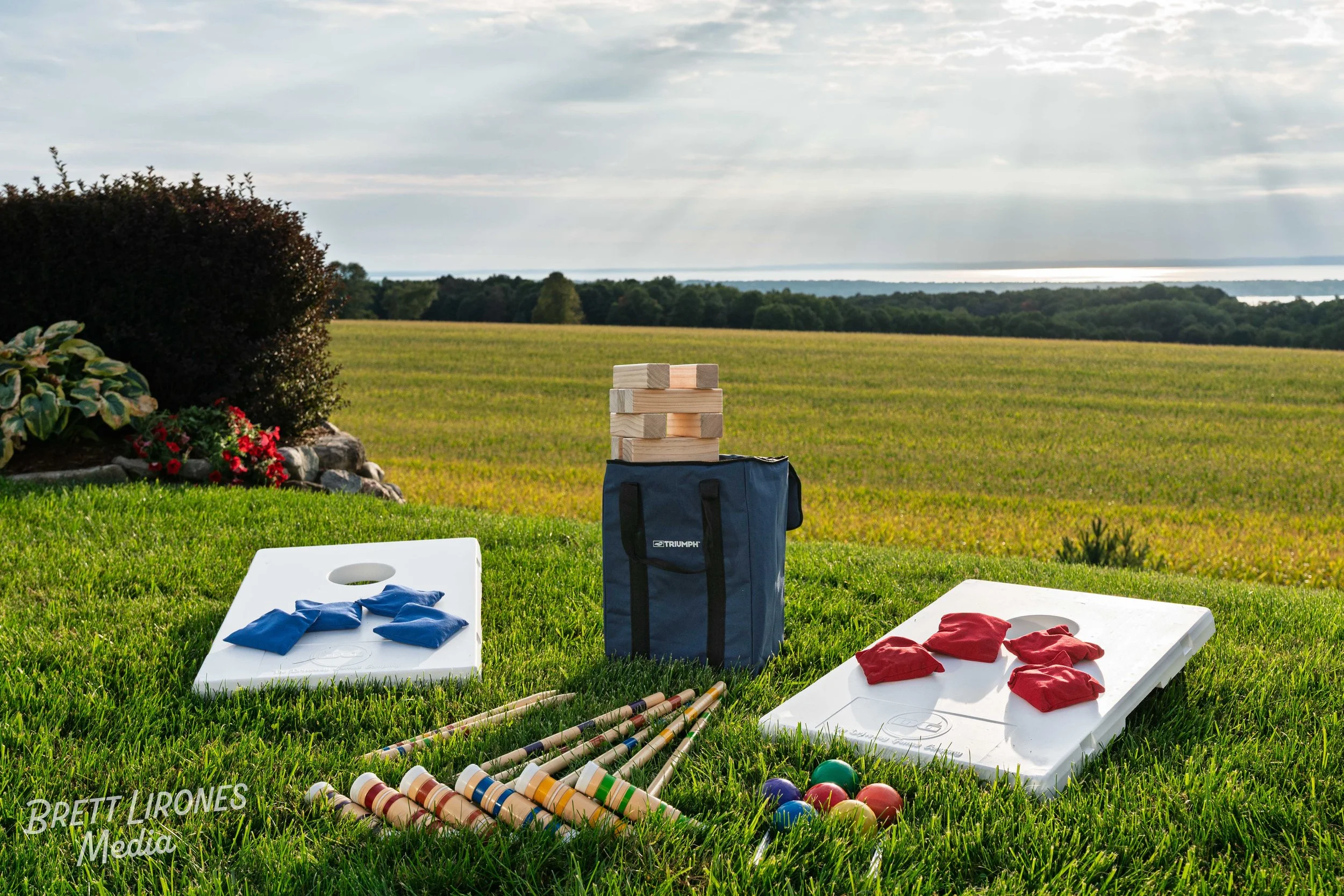 Cornhole game setup on grass with wood blocks, bean bags, and a carrying bag with a scenic field and water in the background.