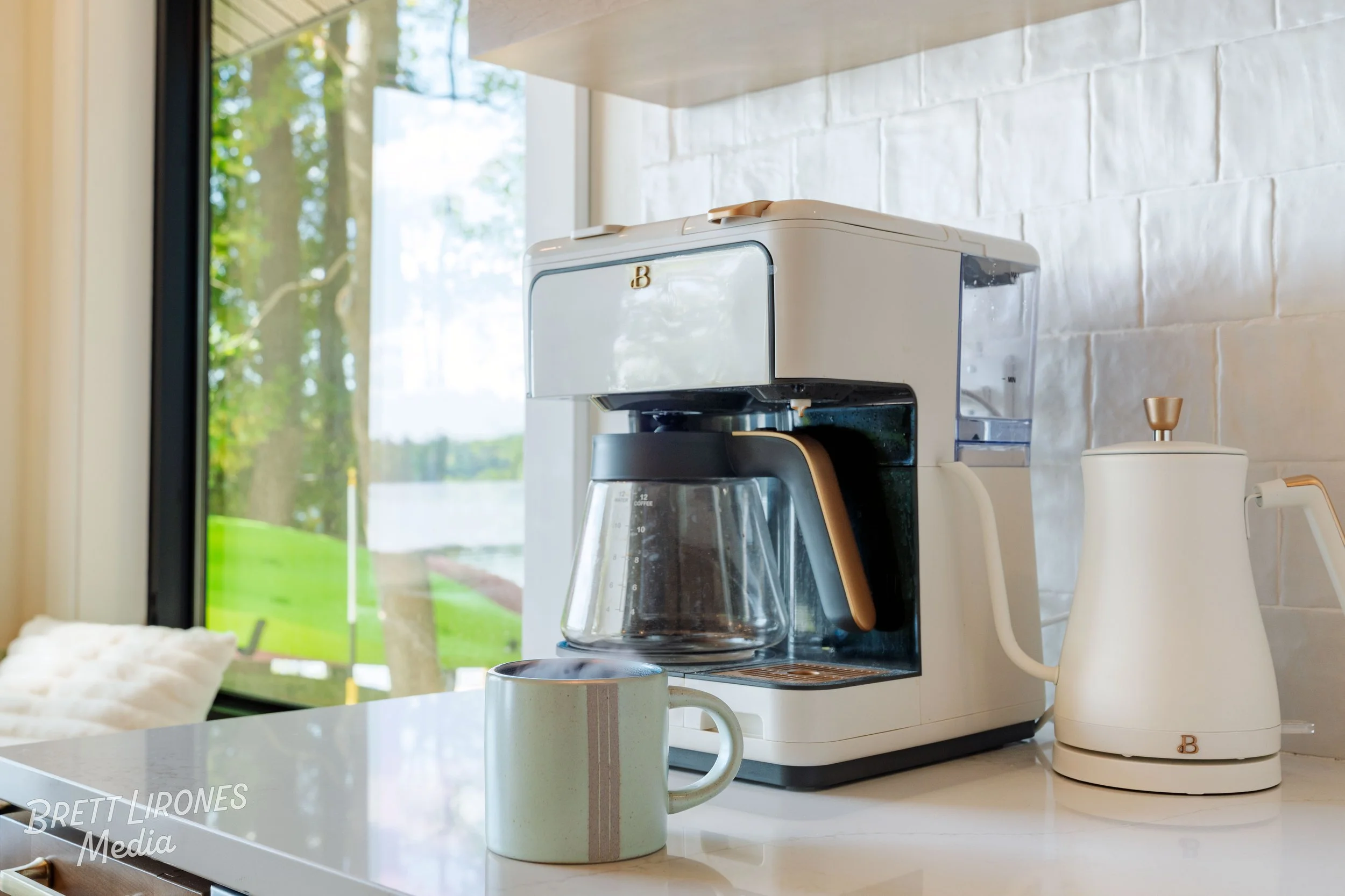 Coffee machine on a kitchen counter with a mug in front and a white kettle to the right, near a window with trees outside.