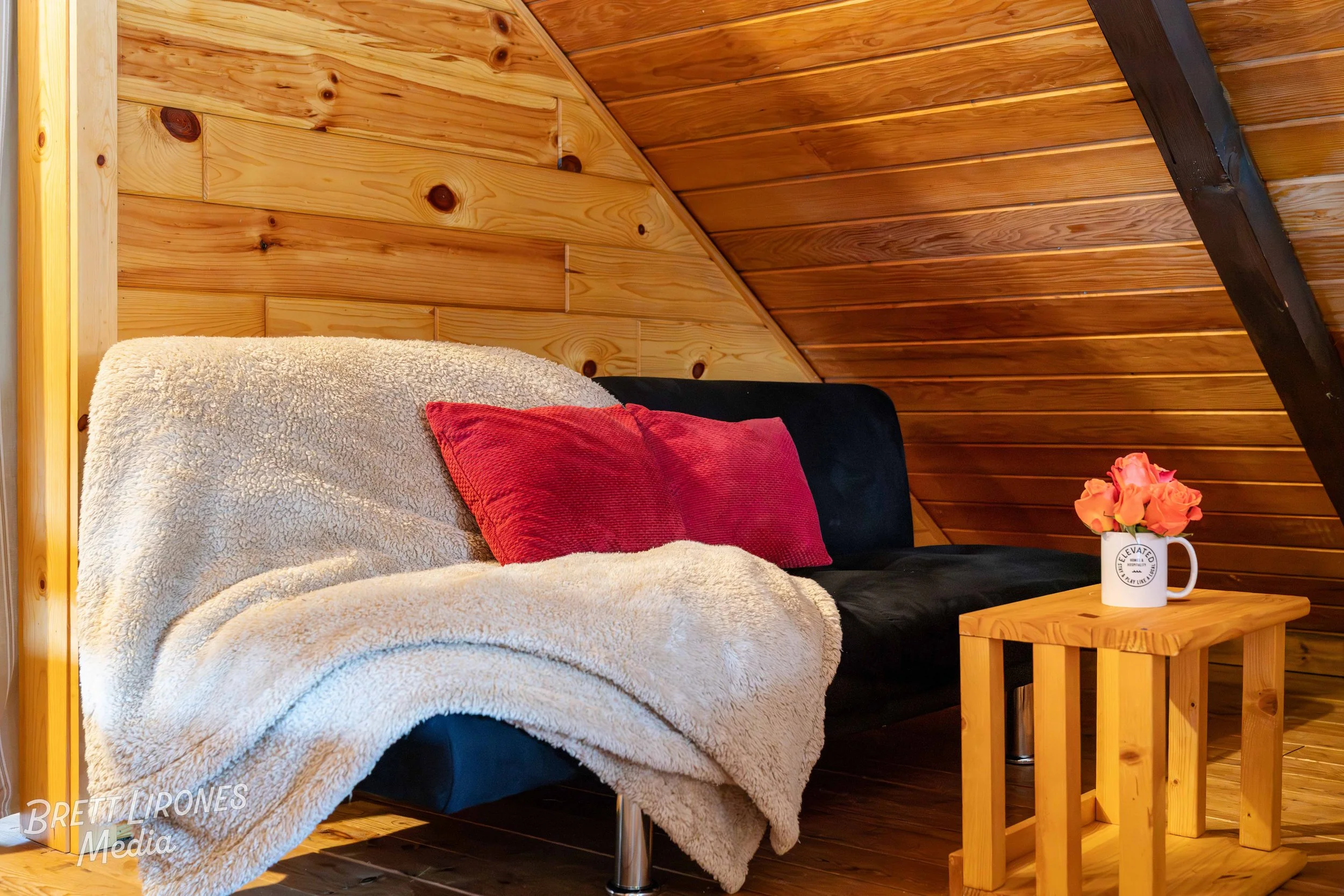 A cozy wooden-paneled room with a black futon covered by a beige fleece blanket and accented with two red pillows. A small wooden side table holds a white mug filled with pink roses, creating a warm inviting atmosphere.
