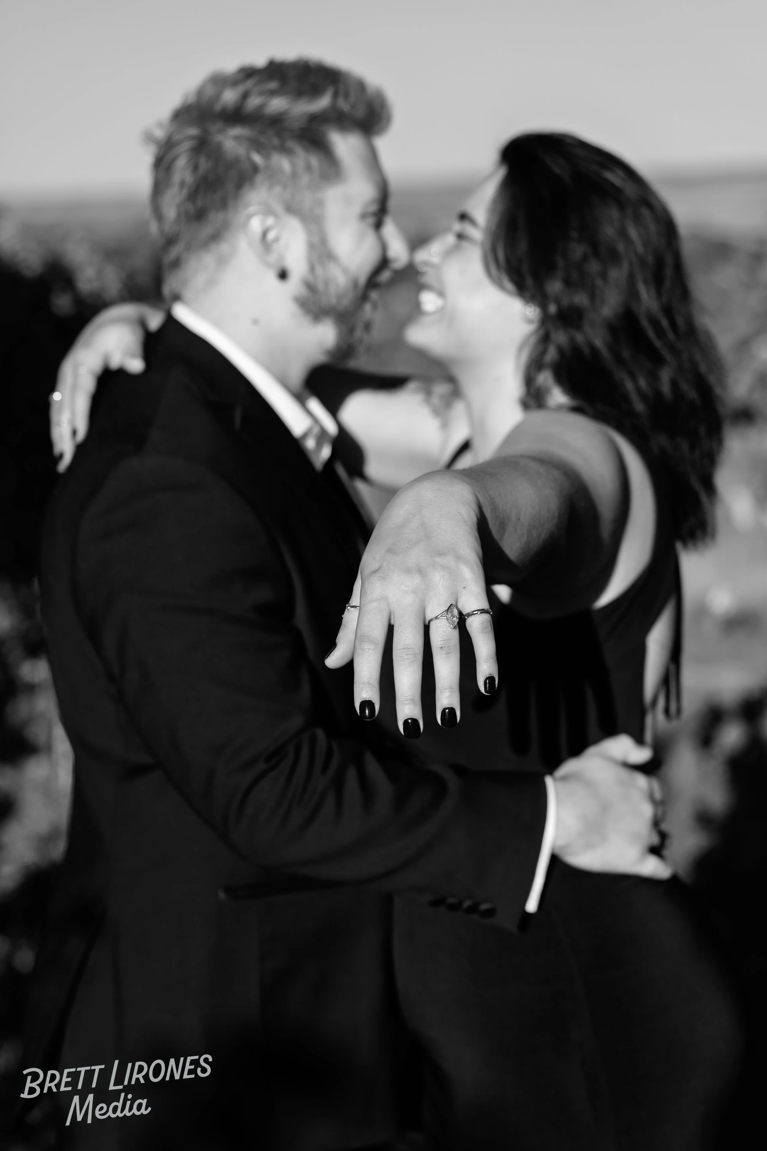 A black and white photo of a couple smiling, embracing, and showing their hands with wedding rings, outdoors with a blurred natural background.