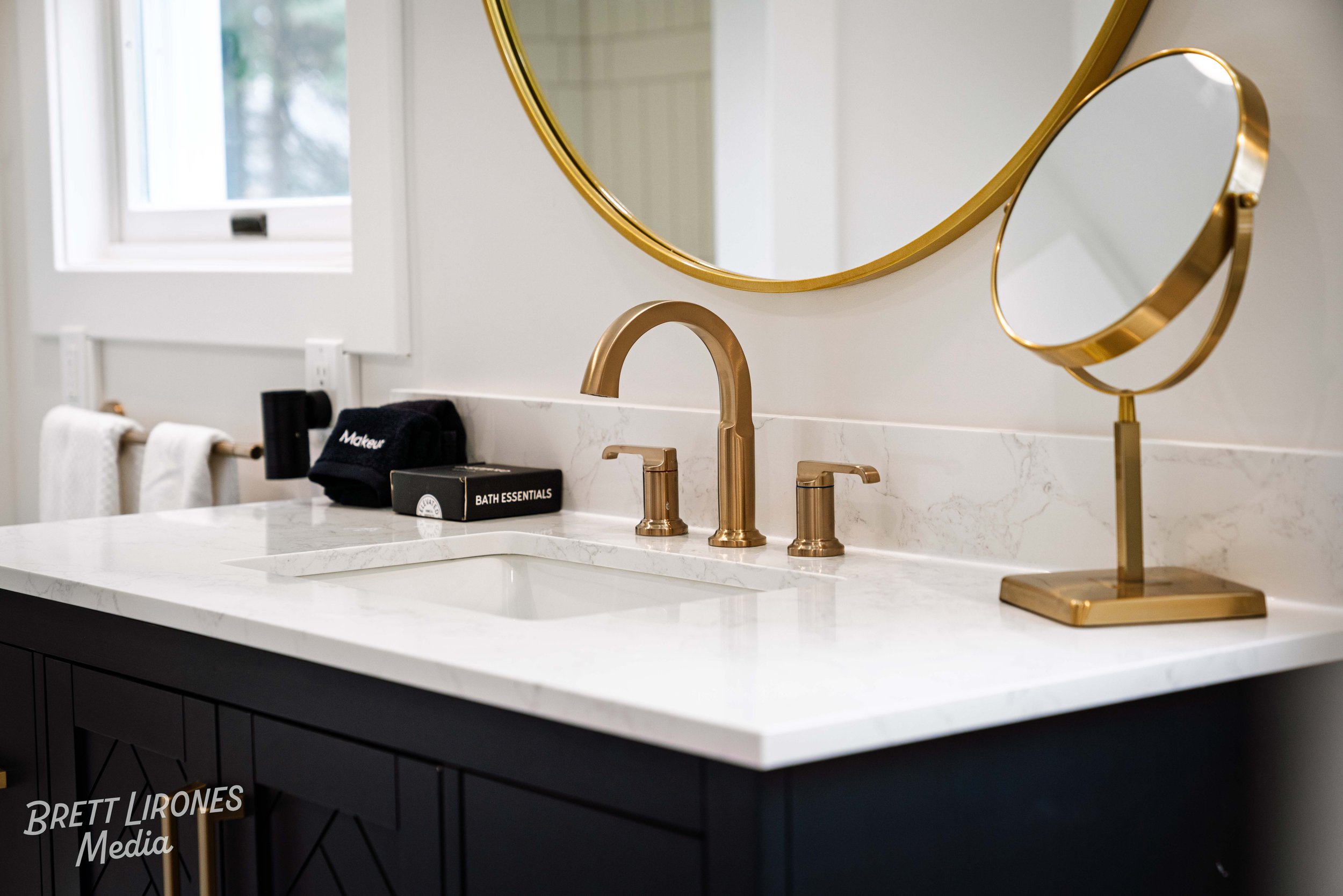 A modern bathroom vanity with a white marble countertop, a gold faucet, and two gold decorative mirrors. There are black towels and bath essentials on the counter, with a window in the background letting in natural light.