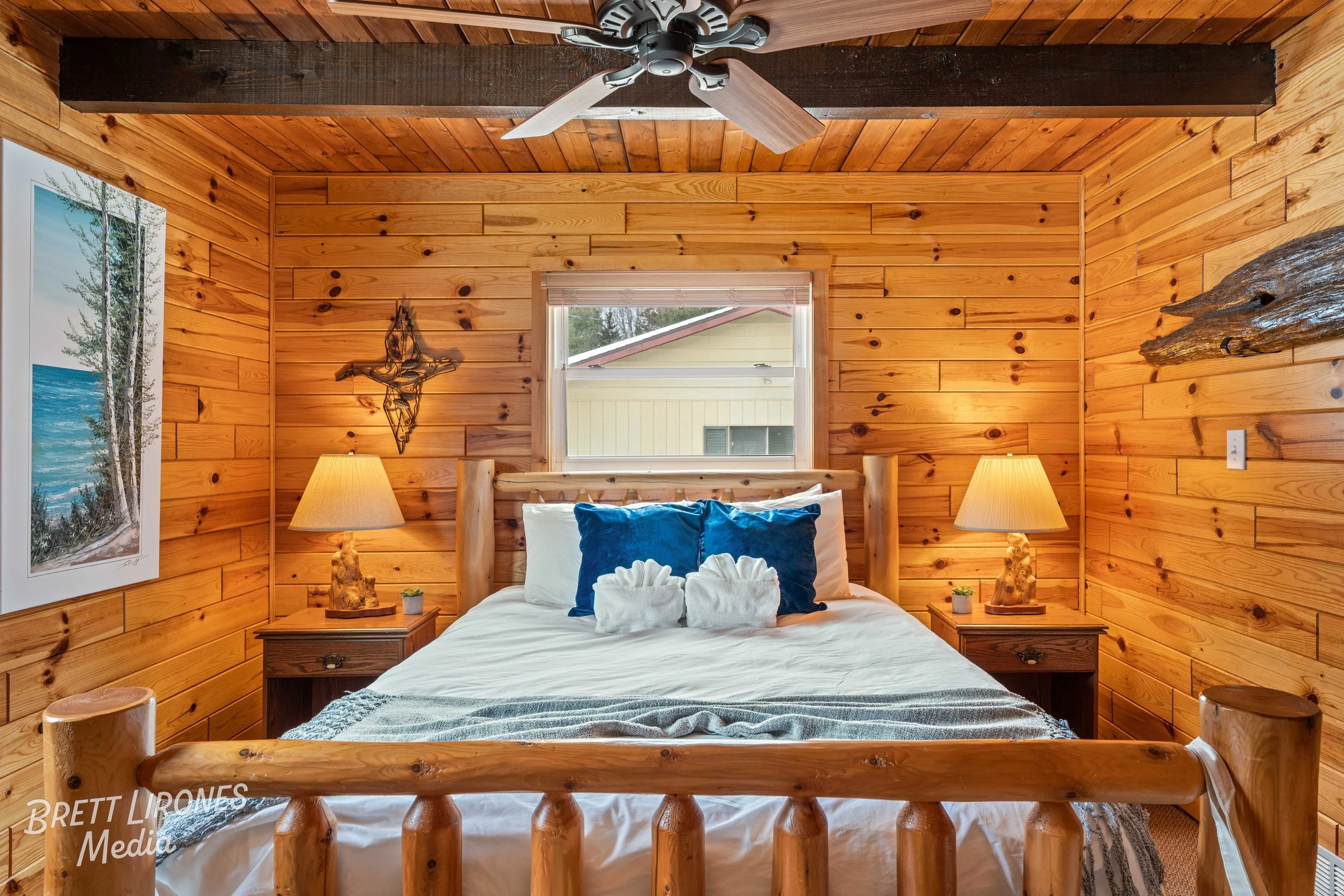 Cozy bedroom with wooden paneled walls, a wooden bed frame, and two matching bedside tables with lamps. The bed has white bedding and blue pillows, with a window above the headboard showing a view of a house and trees outside.