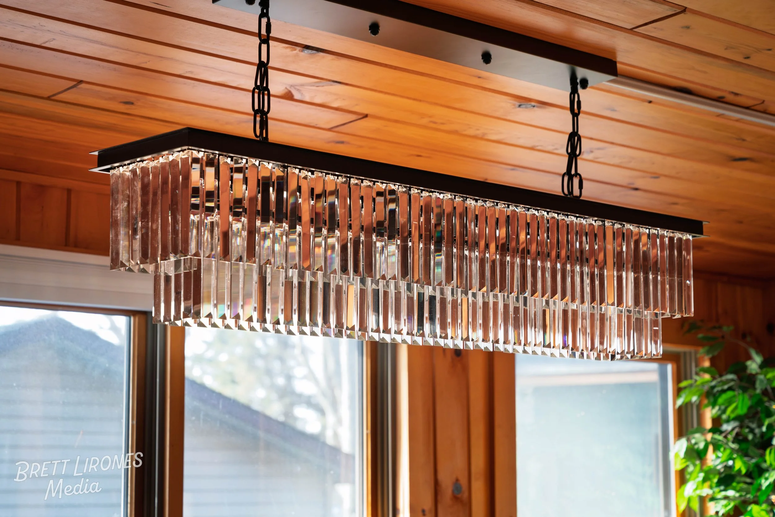 Modern rectangular chandelier with hanging crystal prisms, suspended by black chains from a wooden ceiling inside a room with large windows and wood-paneled walls.