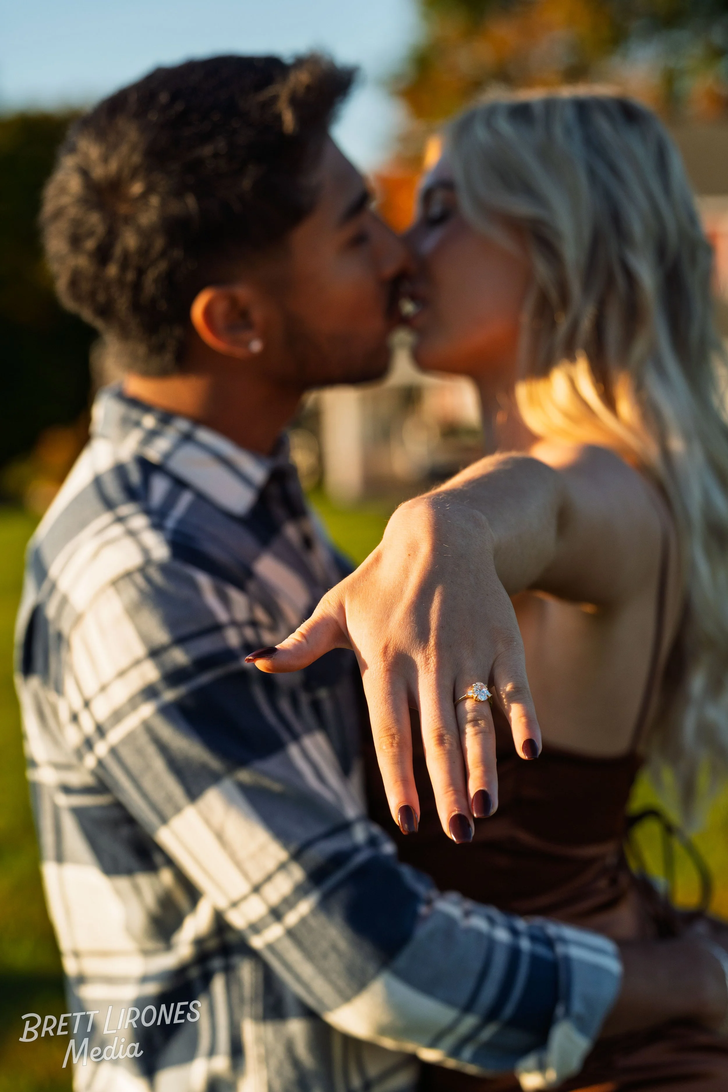 A couple kissing outdoors during golden hour, with the woman showing off a large engagement ring on her finger.