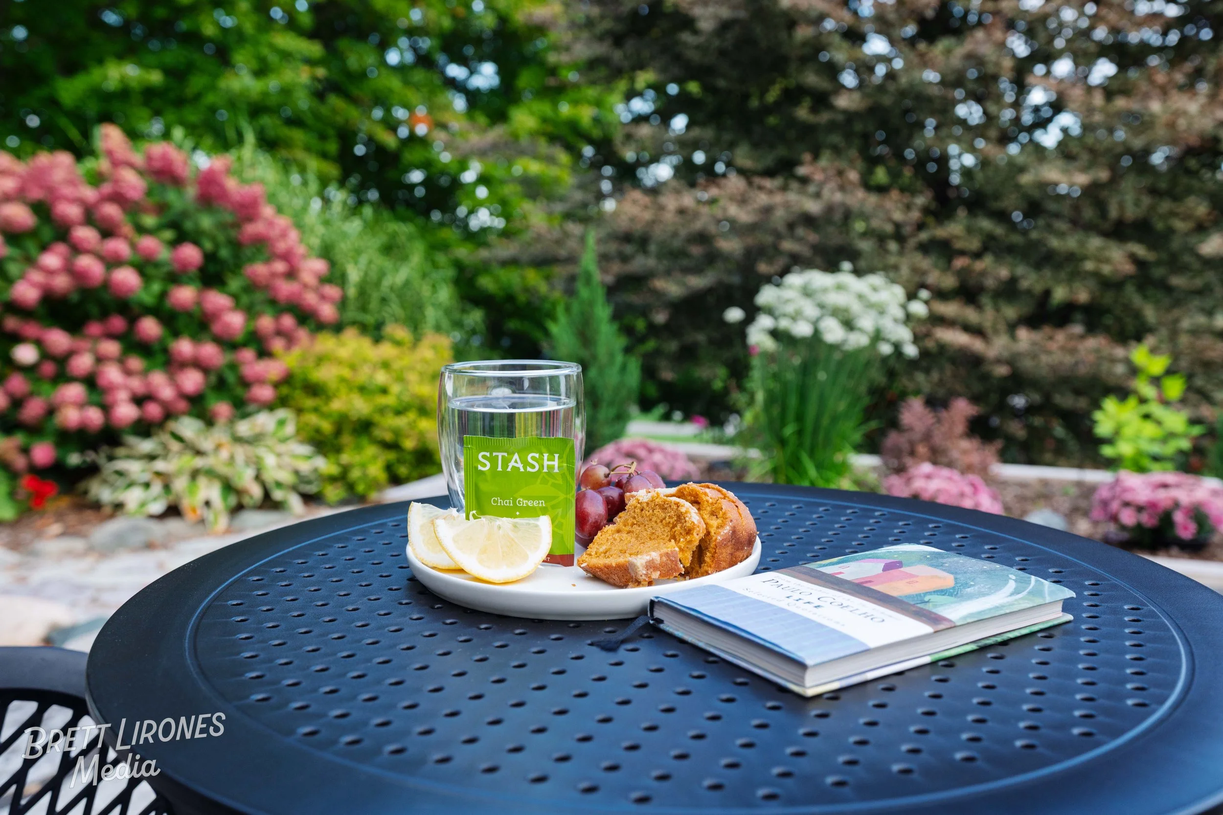 Outside patio table with a glass of water, lemon slices, a slice of pound cake, a bunch of red grapes, and a magazine, with a lush garden full of colorful flowers and trees in the background.