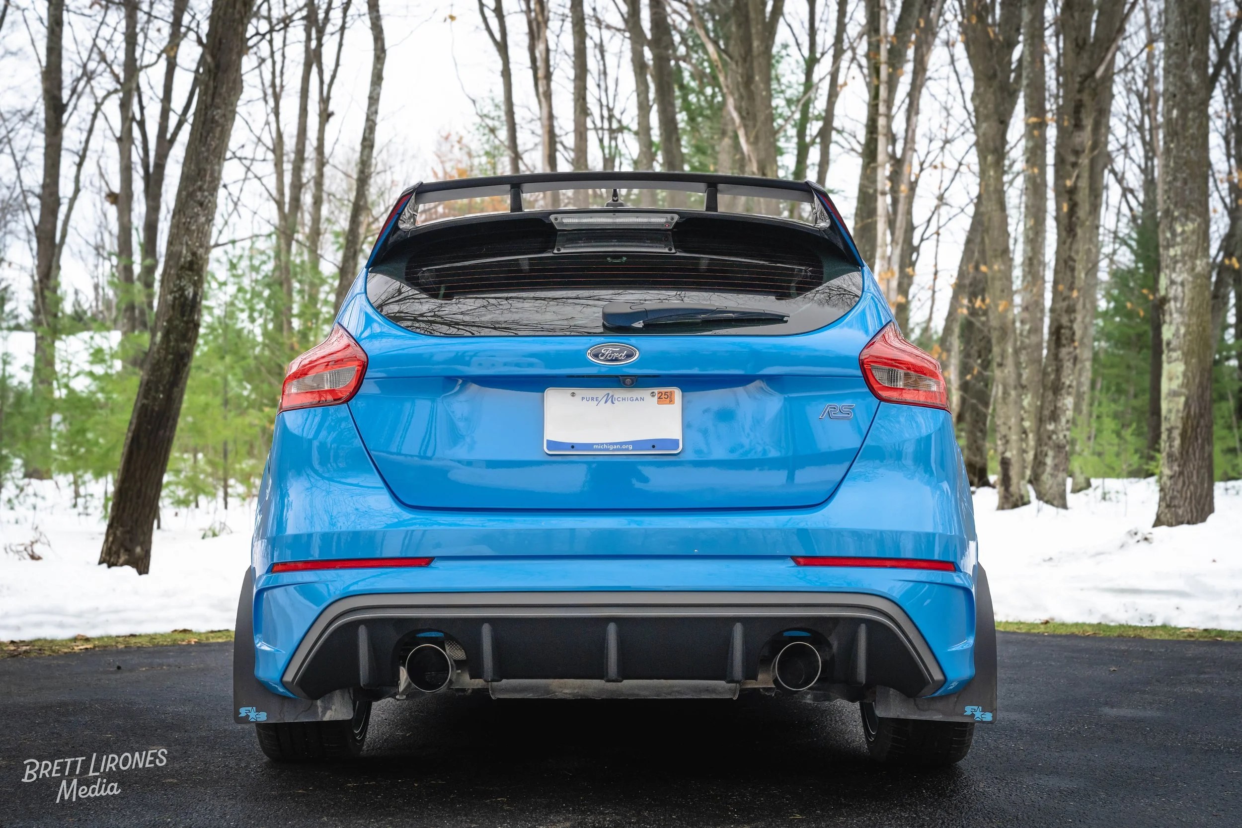 Rear view of a blue Ford RS car with an aftermarket carbon fiber spoiler in a snowy wooded area.