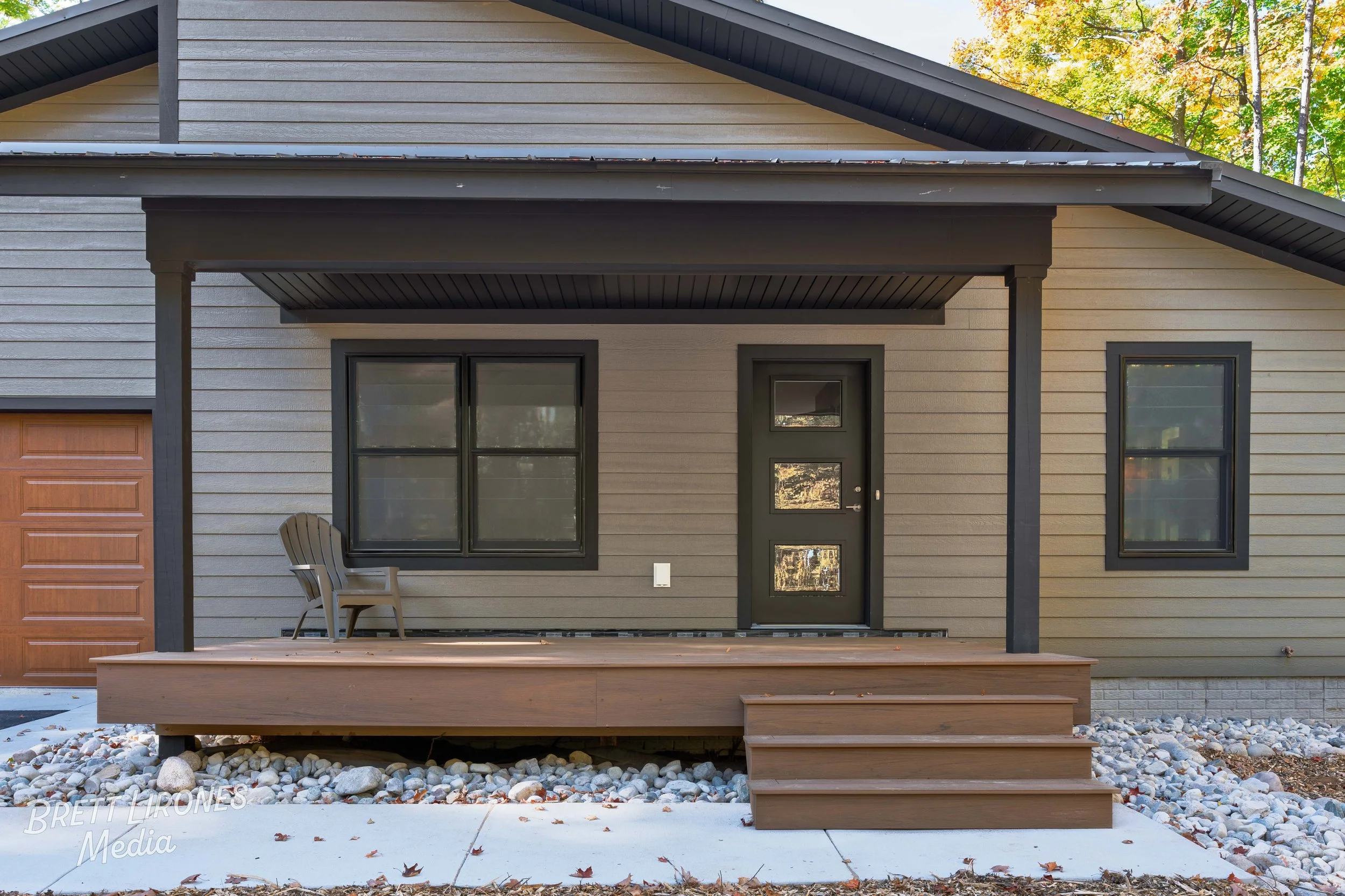Front view of a house with a small porch, black exterior door with three glass panels, two black-framed windows, a brown garage door, a single chair on the porch, and a stepped wooden ramp, with rocks and concrete in the surrounding yard and trees in