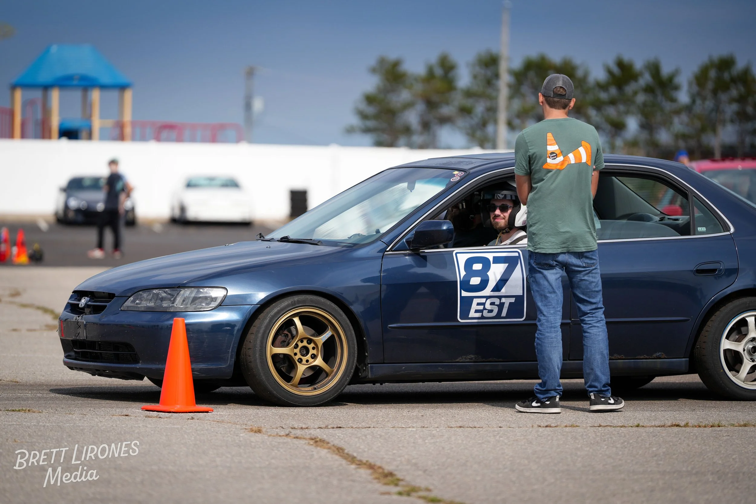 A man in casual clothes and a baseball cap stands outside a navy blue racing car with the number 87 and the word EST on a door sign, at a racing event on a paved lot. The car has gold wheels and is parked behind an orange traffic cone. In the driver'