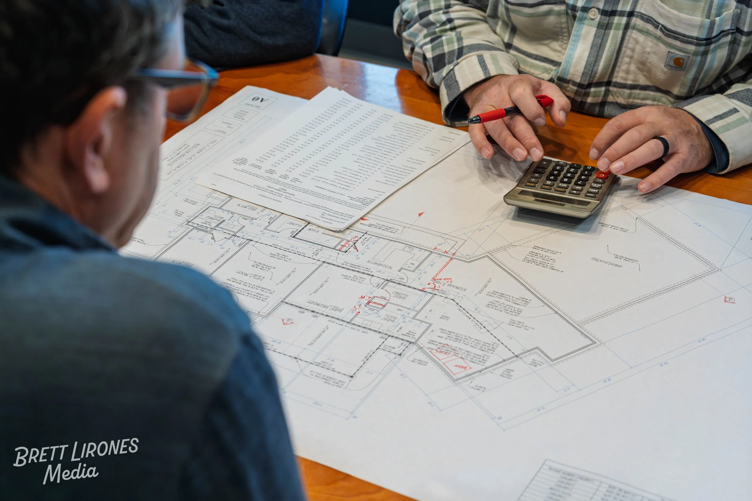 Two men examining architectural blueprints and documents on a wooden table, one of them using a calculator and holding a pen.
