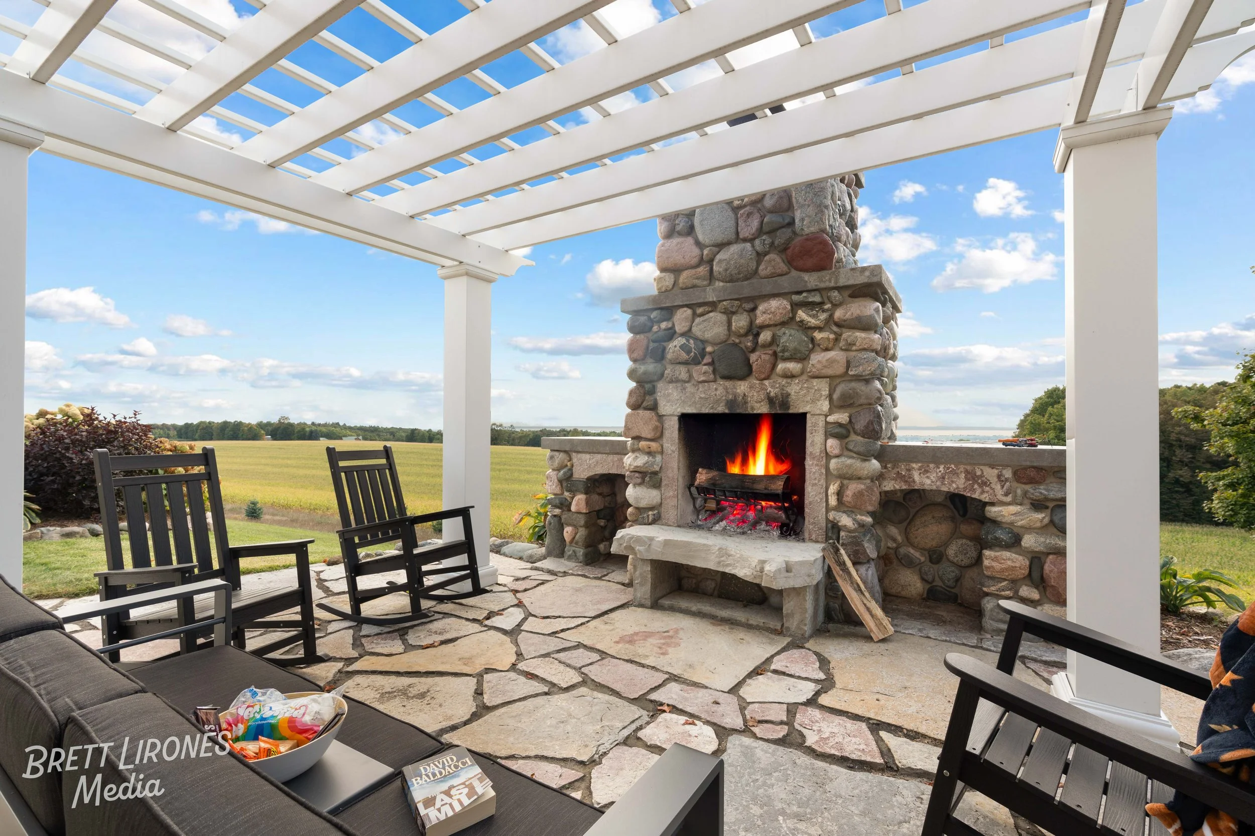An outdoor patio with a stone fireplace burning with a fire, surrounded by black and wooden chairs, a sofa with snacks, and a scenic view of green fields and trees under a partly cloudy sky.