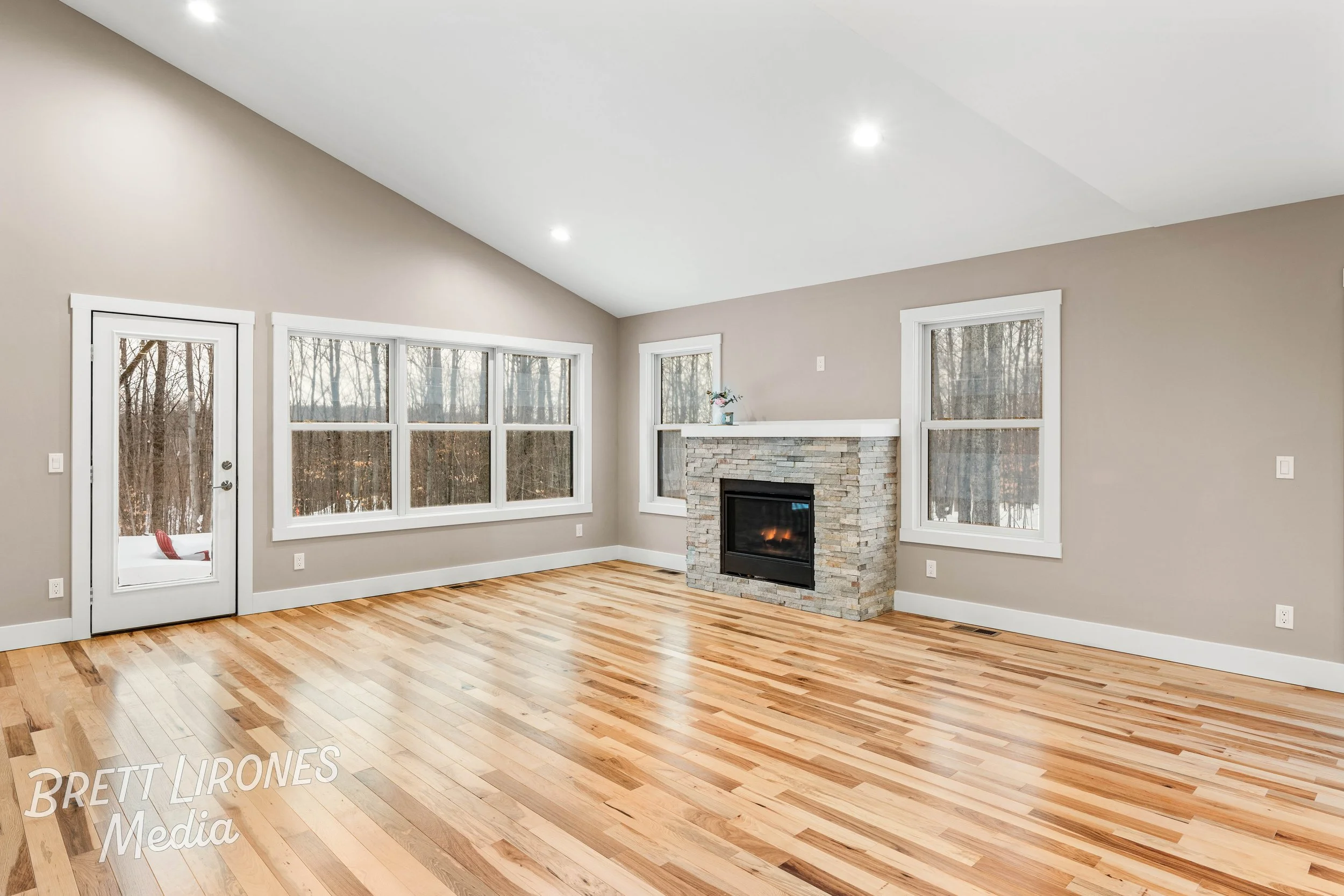 Empty living room with hardwood floors, beige walls, a stone fireplace, large windows, and a glass door leading outside, with a snowy landscape visible through the windows.