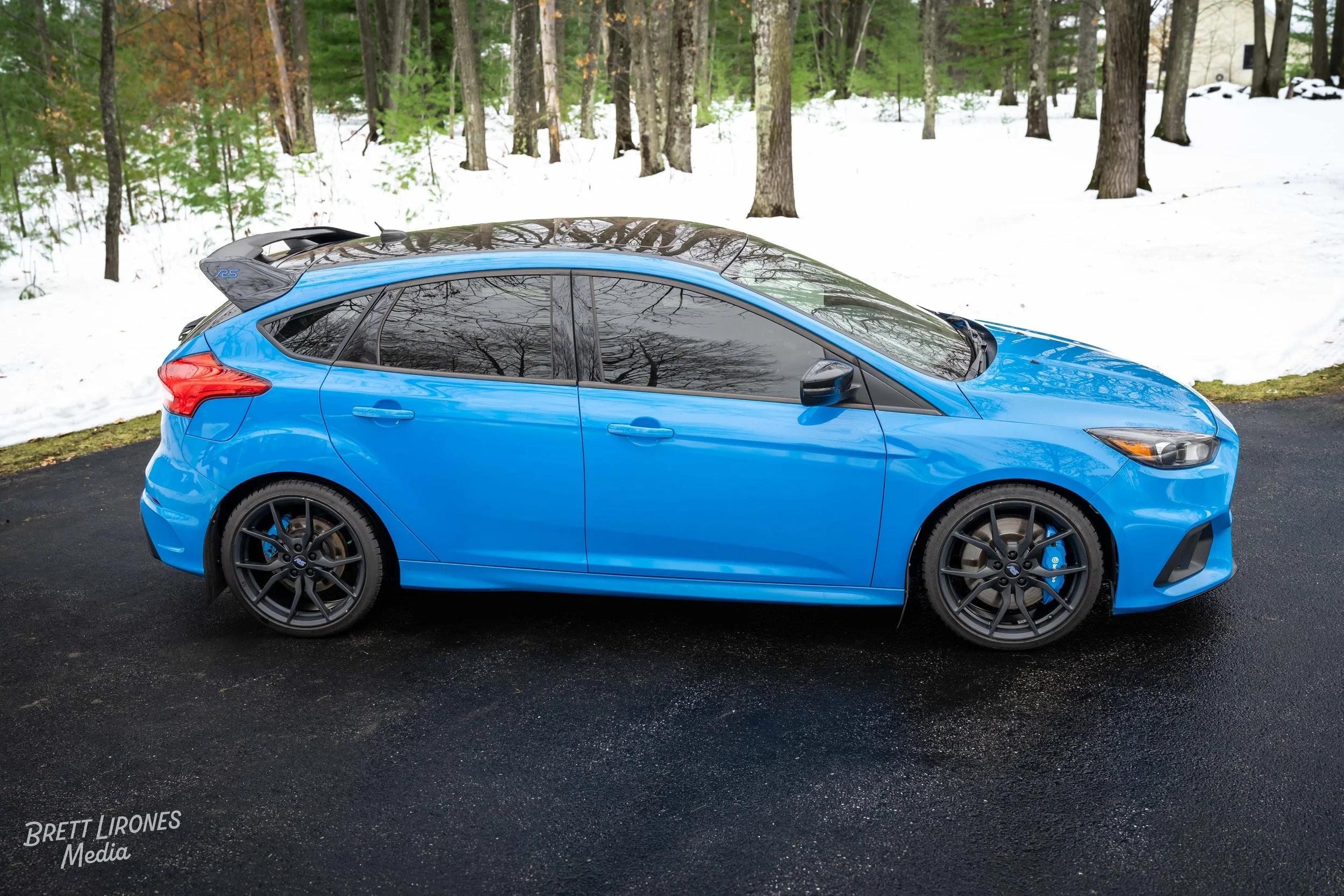 Blue hatchback car parked on a black asphalt driveway with snow and trees in the background.