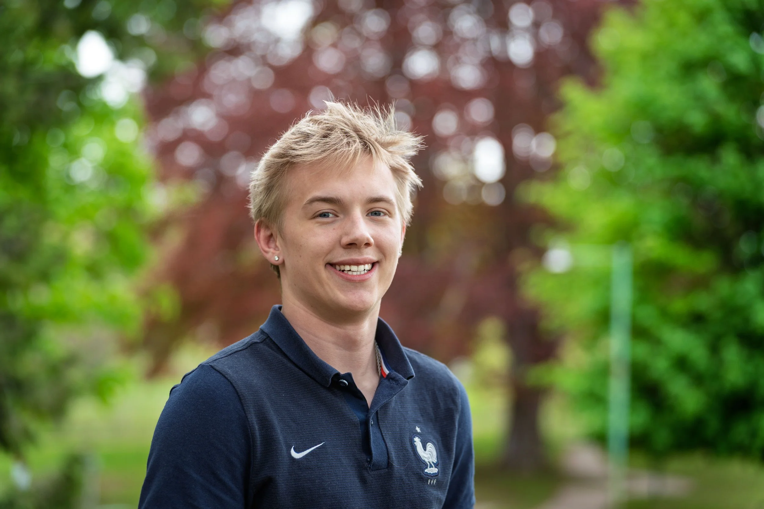 A smiling young man with light hair, wearing a dark blue polo shirt with the French national football team logo, standing outdoors in a park with blurred green and reddish-brown trees in the background.