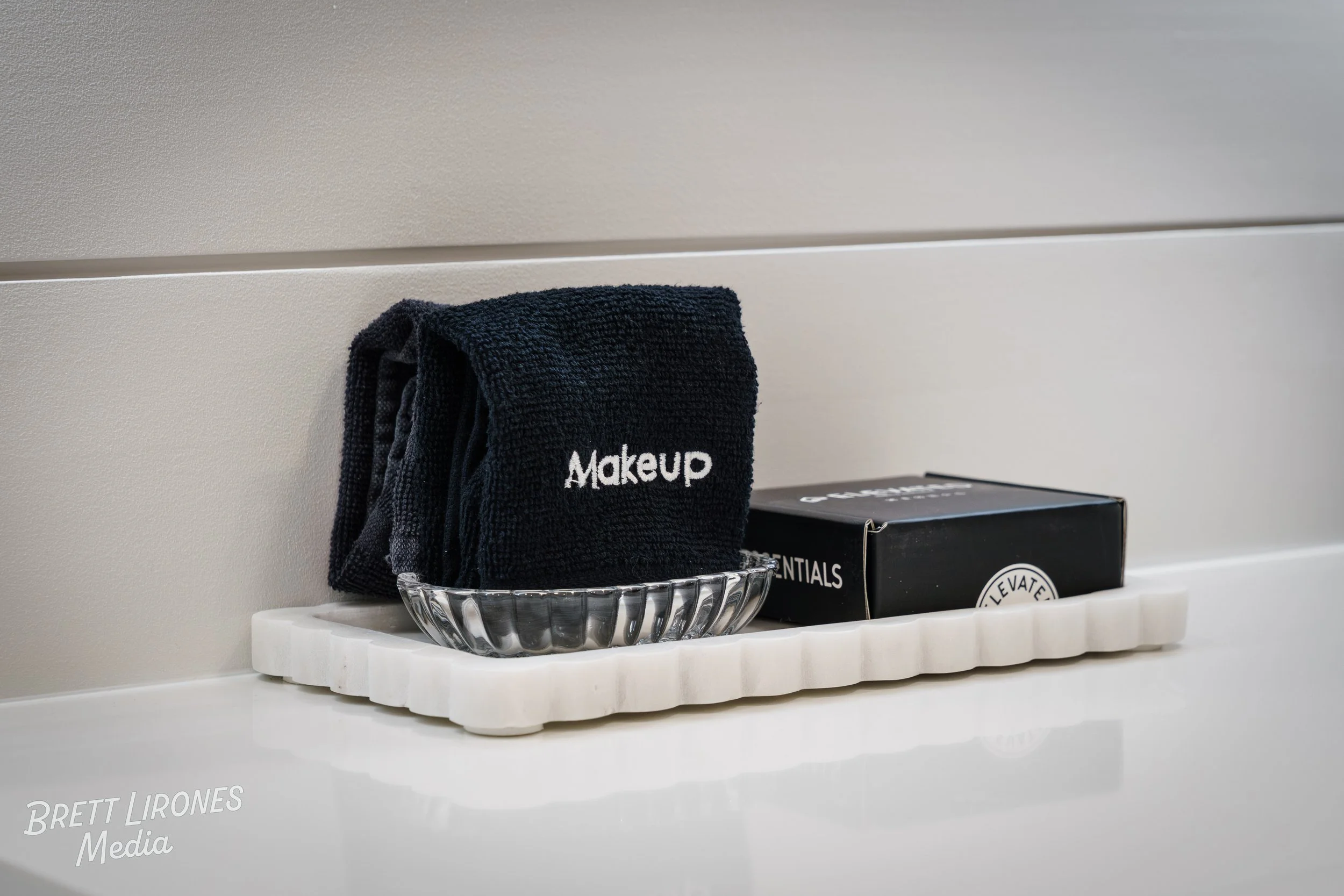 A black towel with white text 'Makeup' hanging on a clear glass bowl, placed on a white marble countertop, with a black box labeled 'Premium Essentials' beside it, against a plain grey wall.