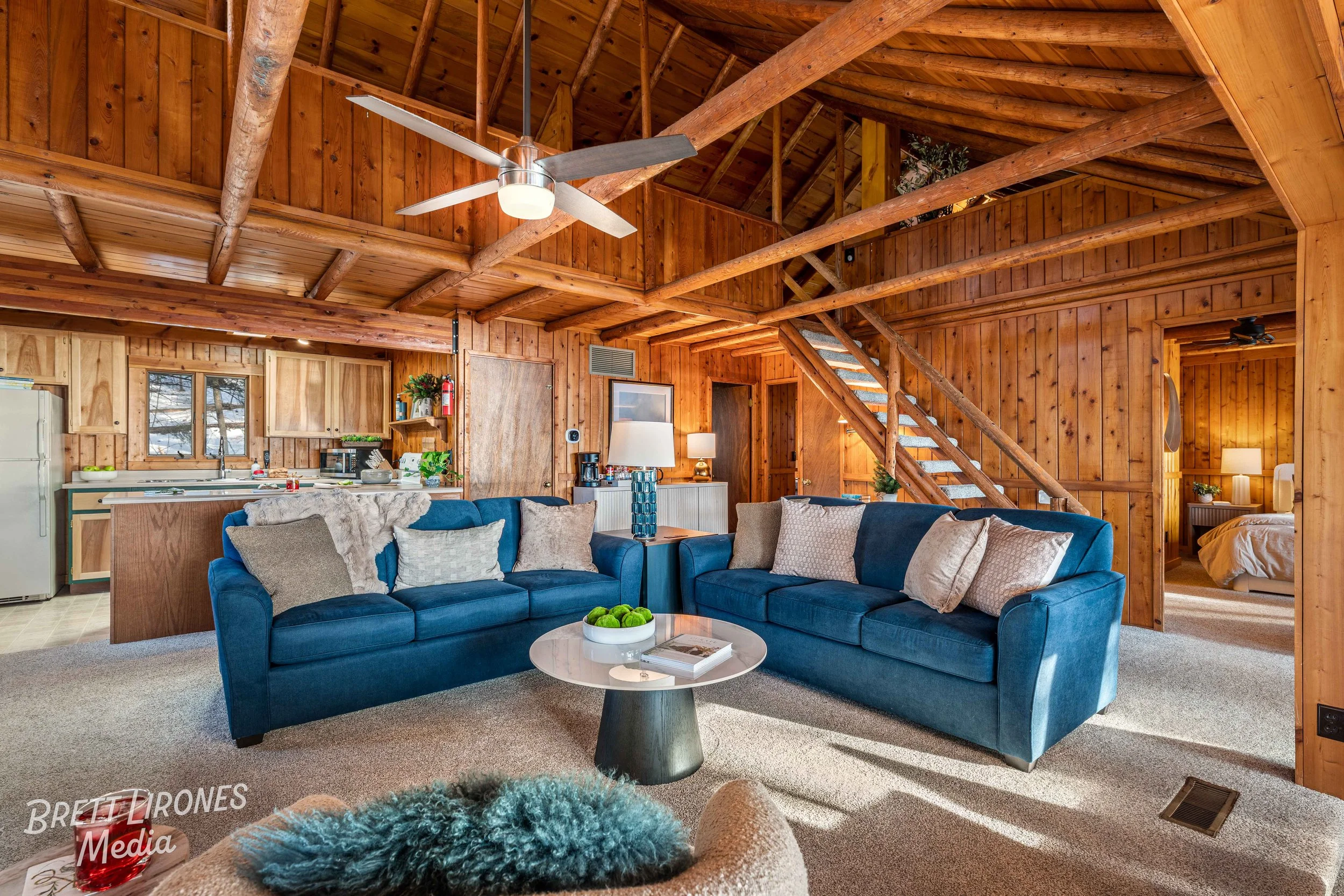 Interior of a rustic wooden cabin living room with blue sofas, a round coffee table, and a staircase leading up to a loft area.