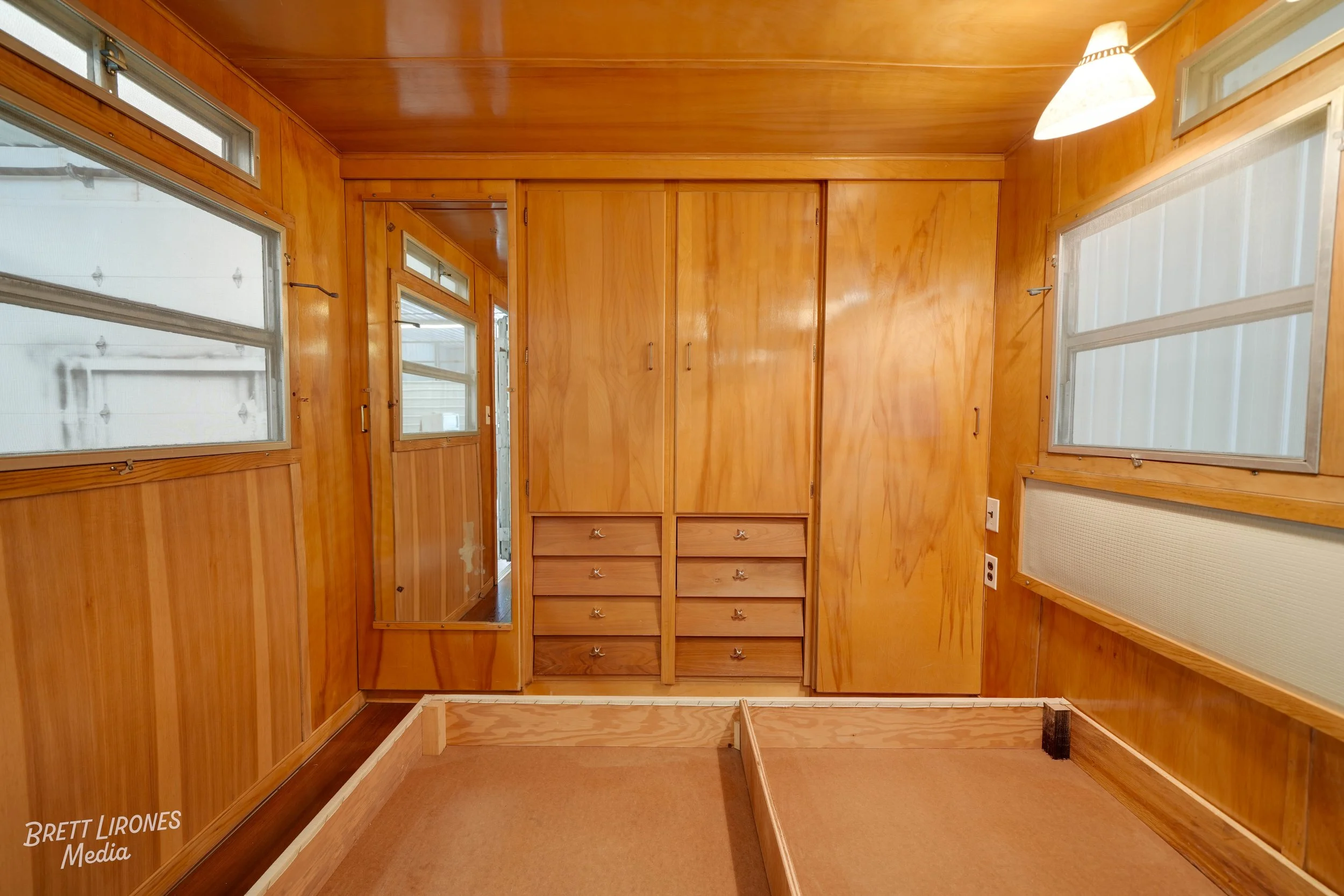 Empty room with wooden walls, built-in closets, some drawers, and windows, possibly part of a house or cabin.