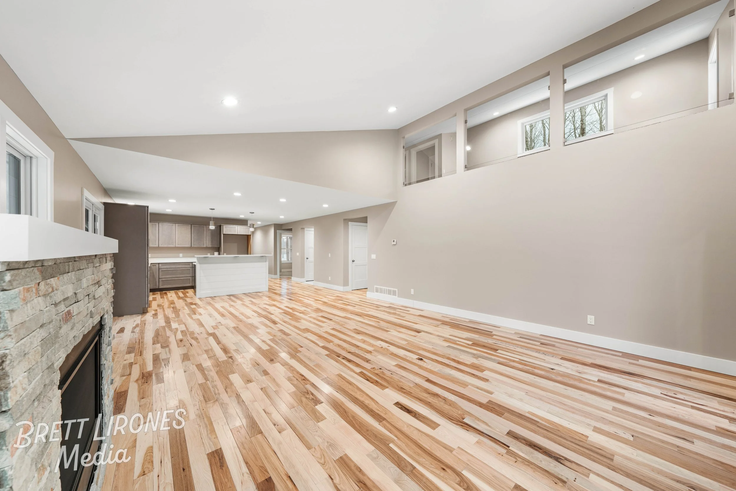 Interior view of an empty living room with hardwood floors, beige walls, a stone fireplace, and a modern kitchen in the background.