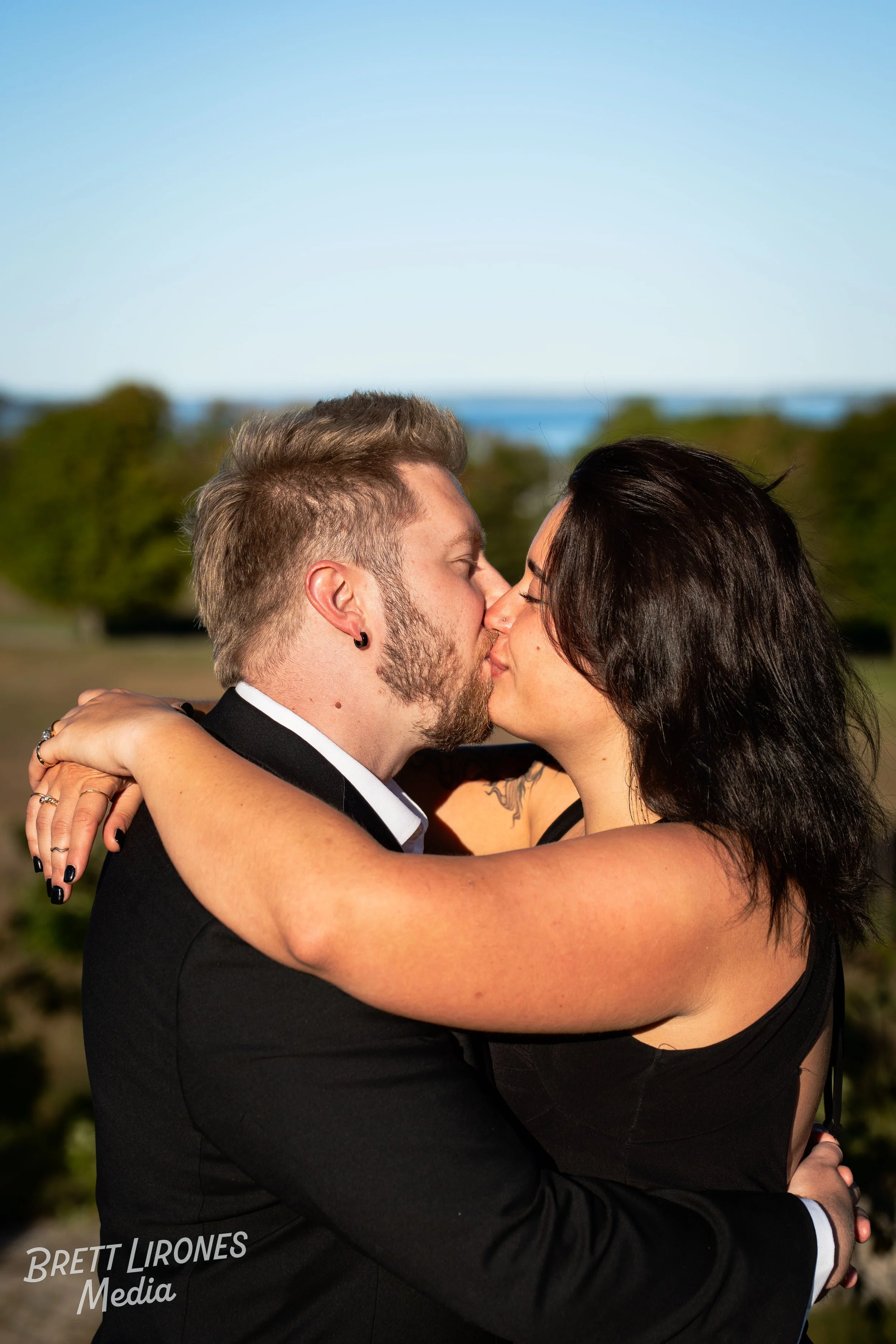 A couple kissing outdoors during daytime, with the man wearing a black suit and the woman in black attire, against a natural background with trees and blue sky.